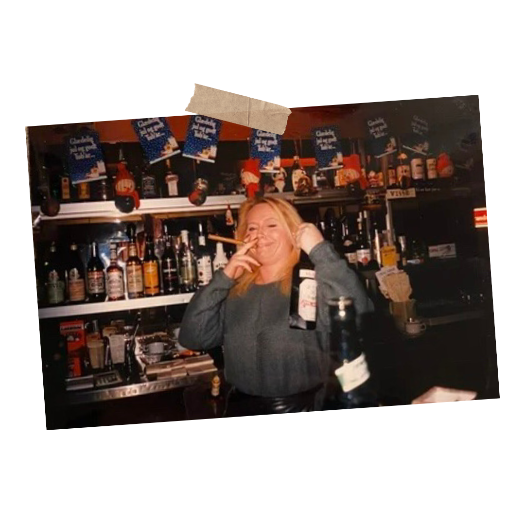 Hende bag bartender, smilende med cigaret i hånden, bag bar med alkoholflasker og julepynt.