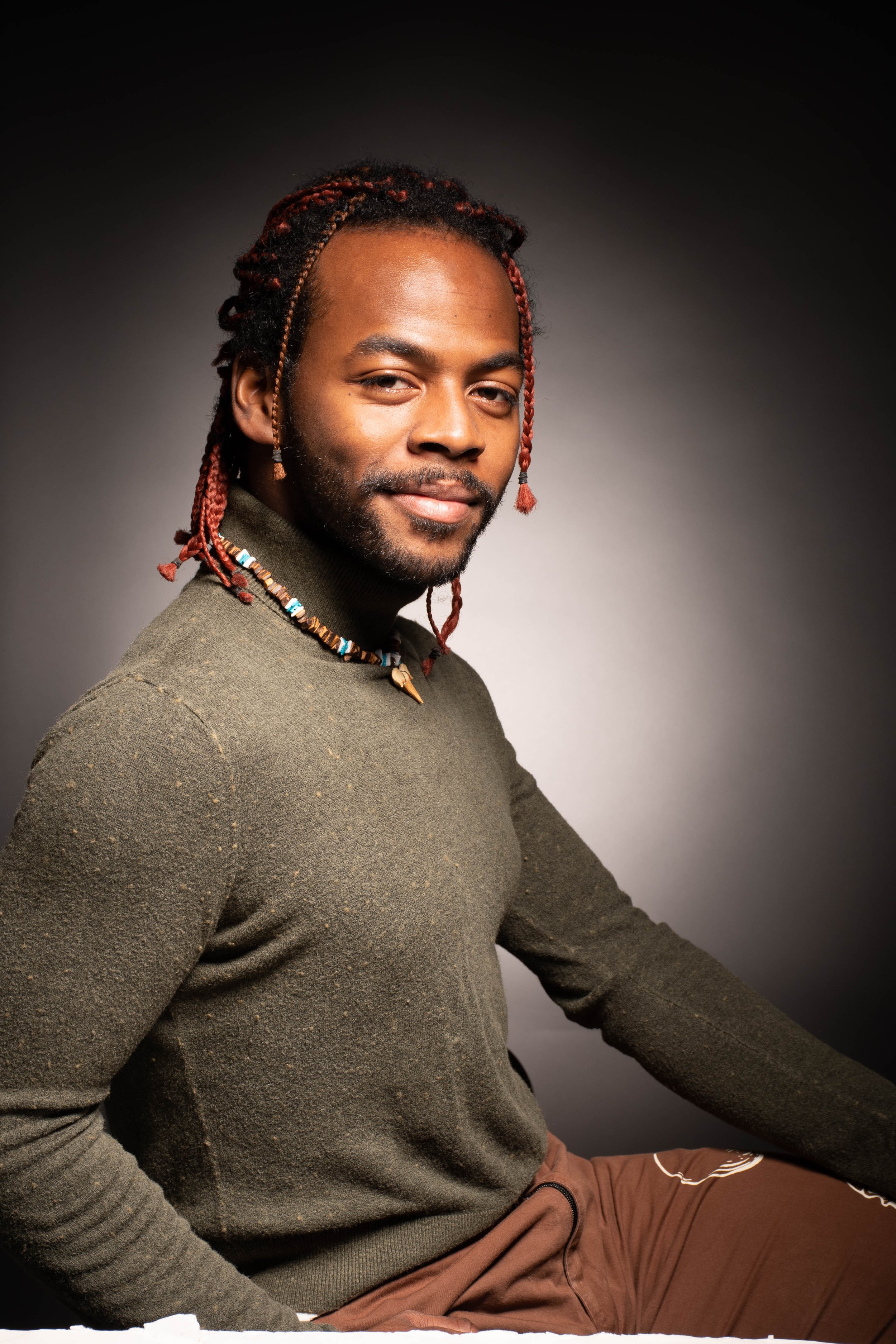Portrait of a man with braided hair and a beard, wearing a beaded necklace and a green sweater, sitting against a plain dark background.