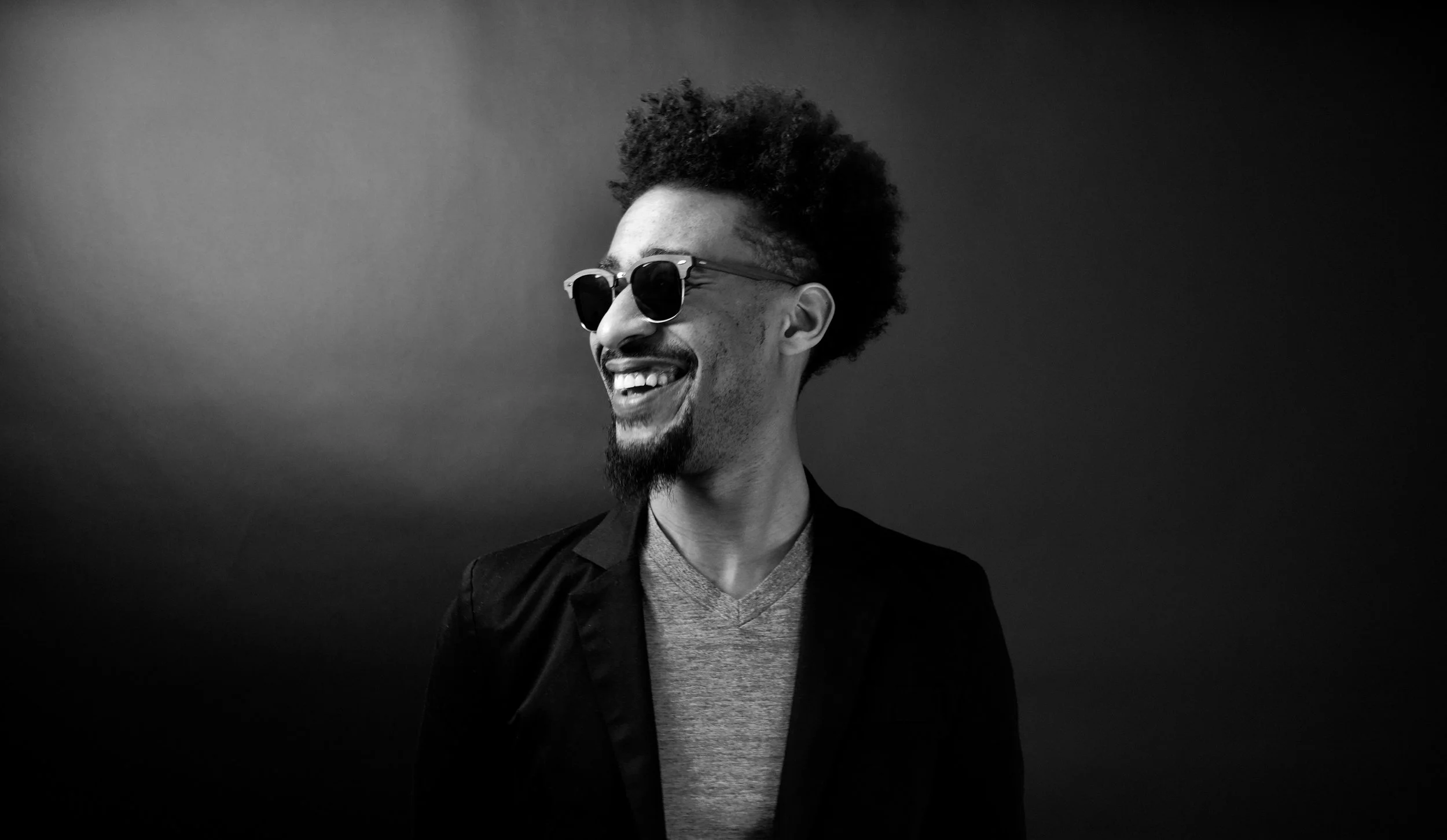 Smiling man with sunglasses, beard, and afro hairstyle on a dark background.