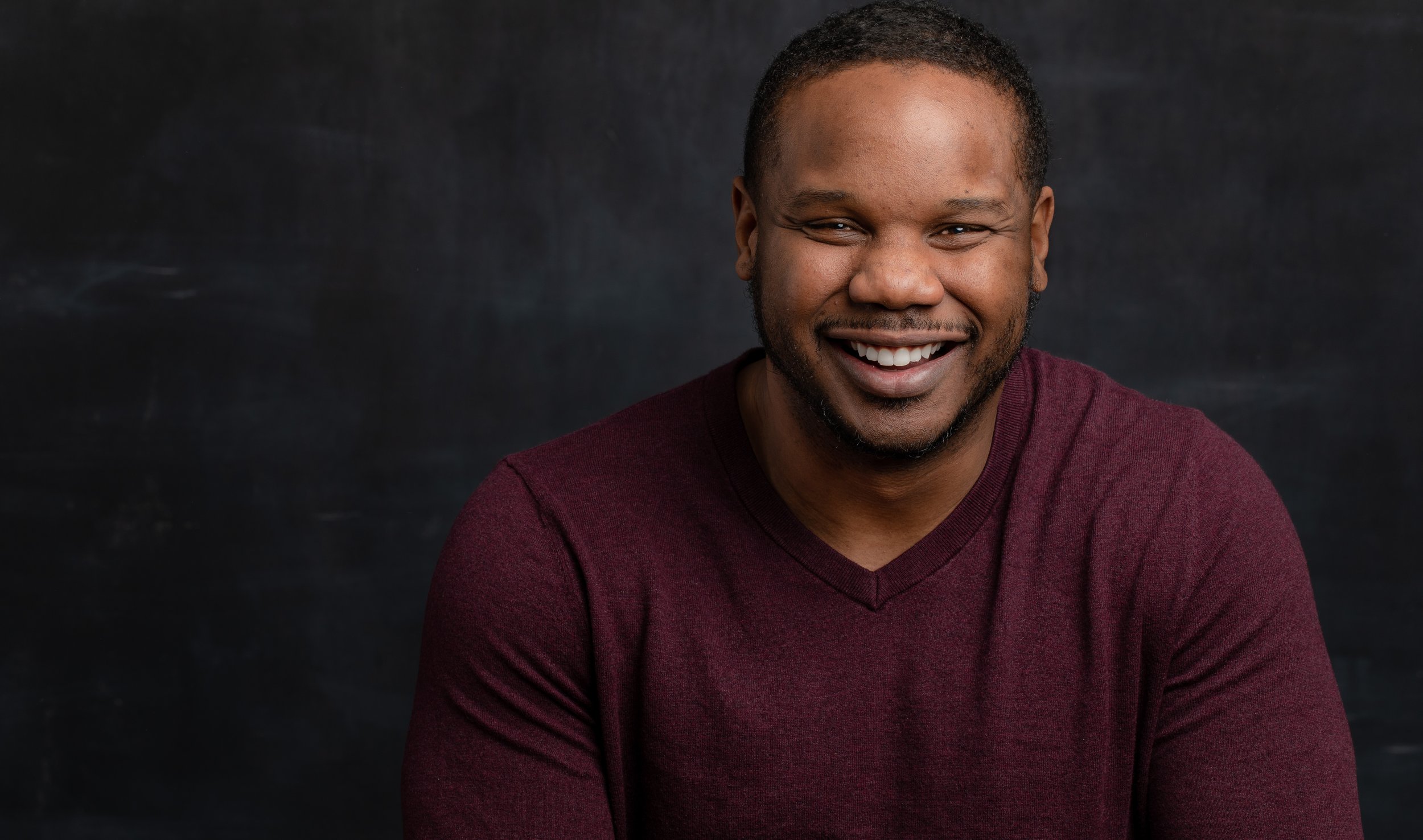 Smiling man with short hair and a burgundy sweater against a dark background.