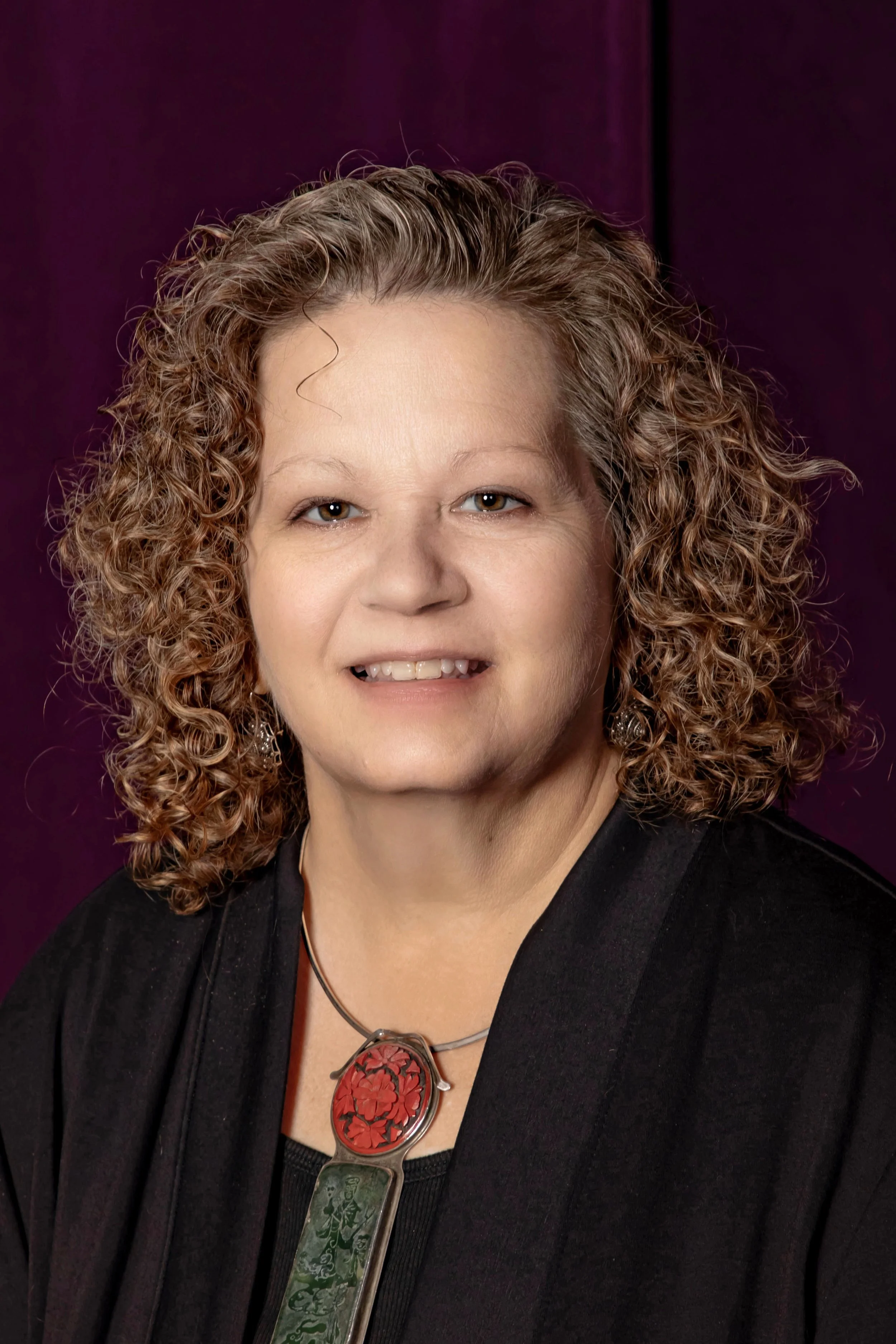 A woman with curly, shoulder-length hair and earrings, wearing a black top and a distinctive pendant necklace, posing against a dark purple background.