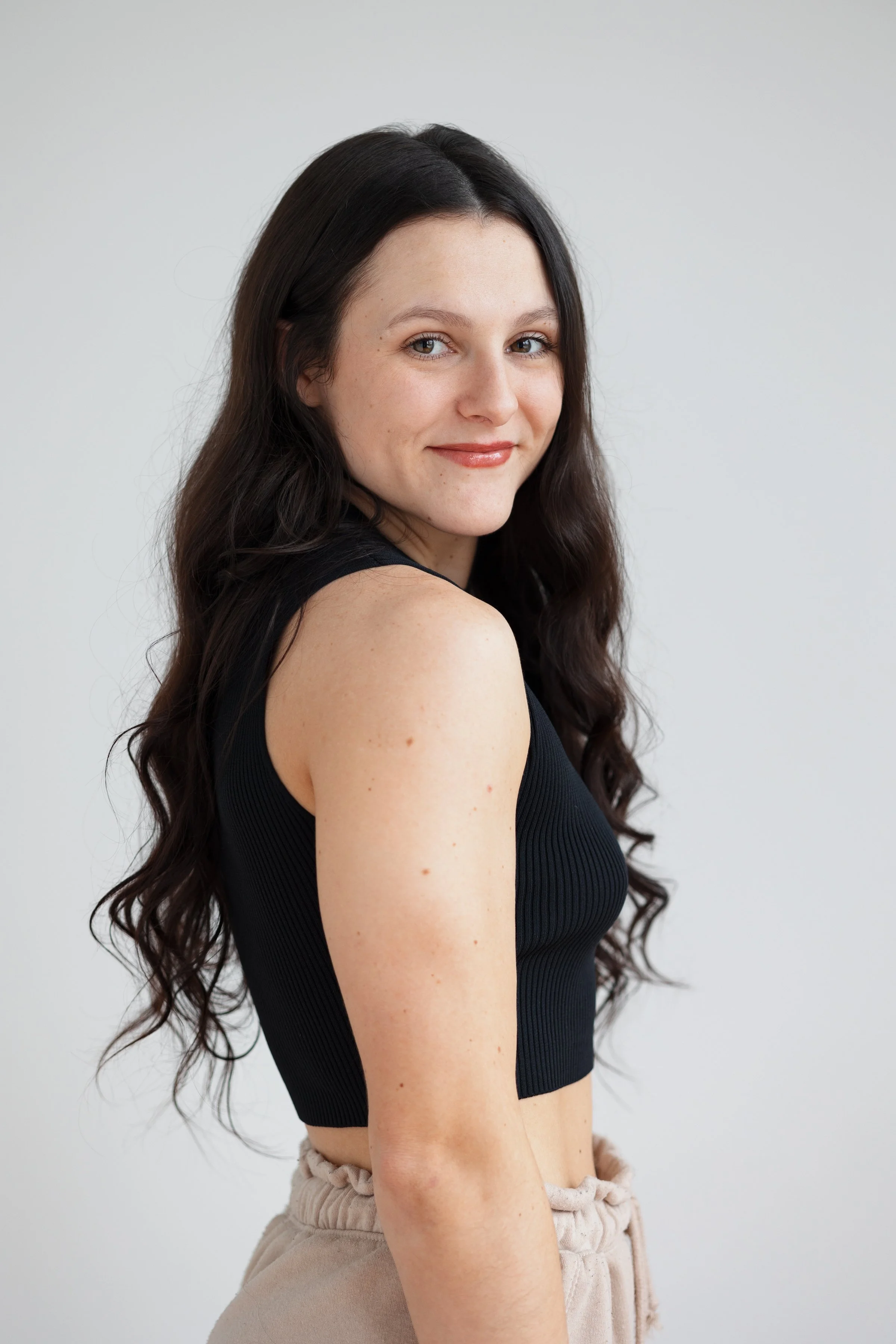 A woman with long dark hair, wearing a sleeveless black crop top and beige sweatpants, smiling softly against a plain light background.