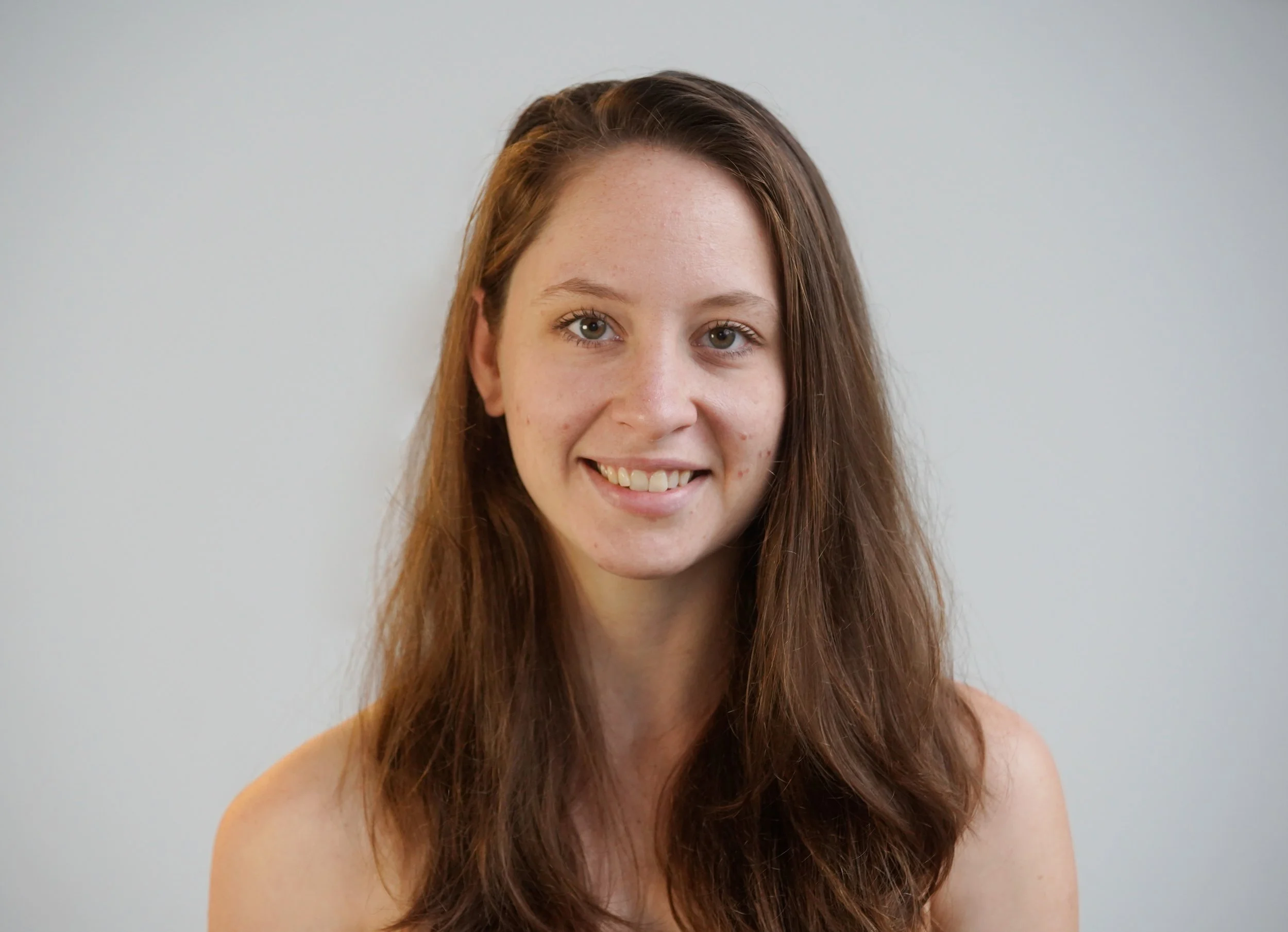 Portrait of a young woman with long, wavy brown hair, smiling, against a plain white background.