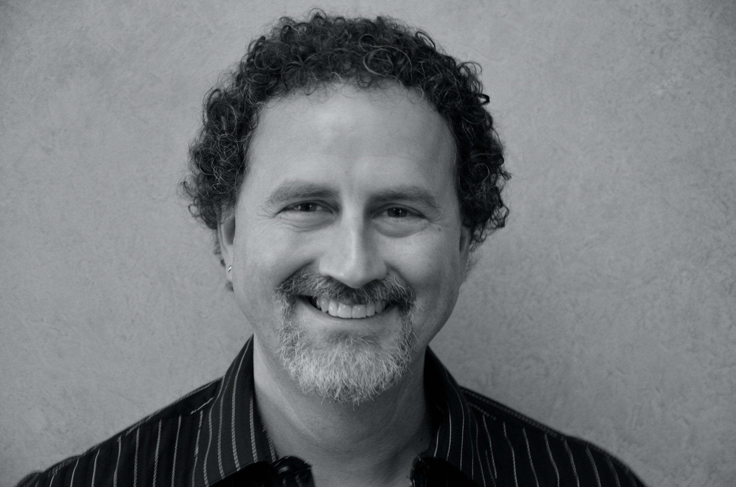 Black and white portrait of a middle-aged man with curly hair, a beard, and a mustache, smiling in front of a plain wall, wearing a striped shirt.