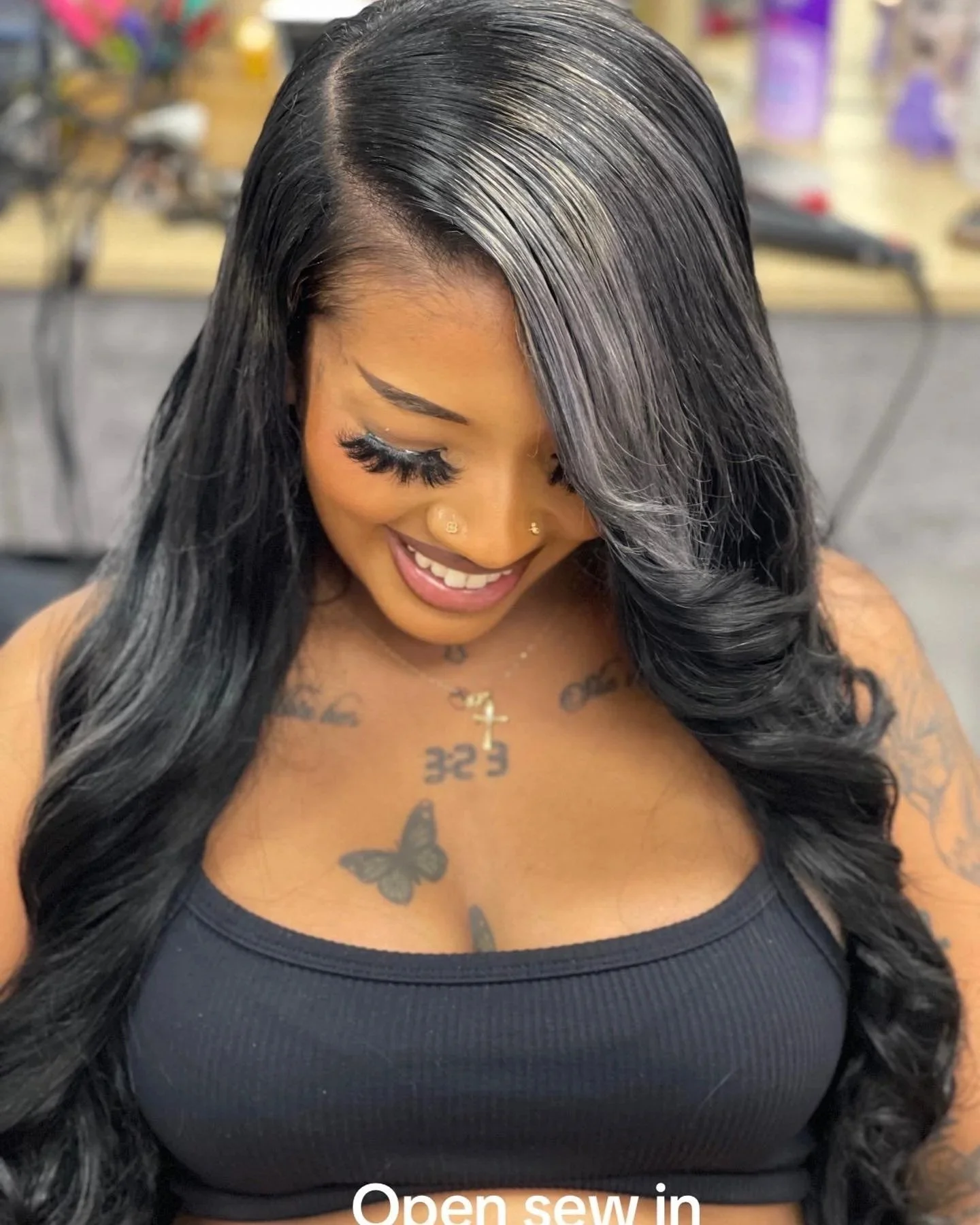 Close-up of a woman with long curly black hair, smiling, wearing a black top, with tattoos on her chest, multiple facial piercings, and heavy makeup.