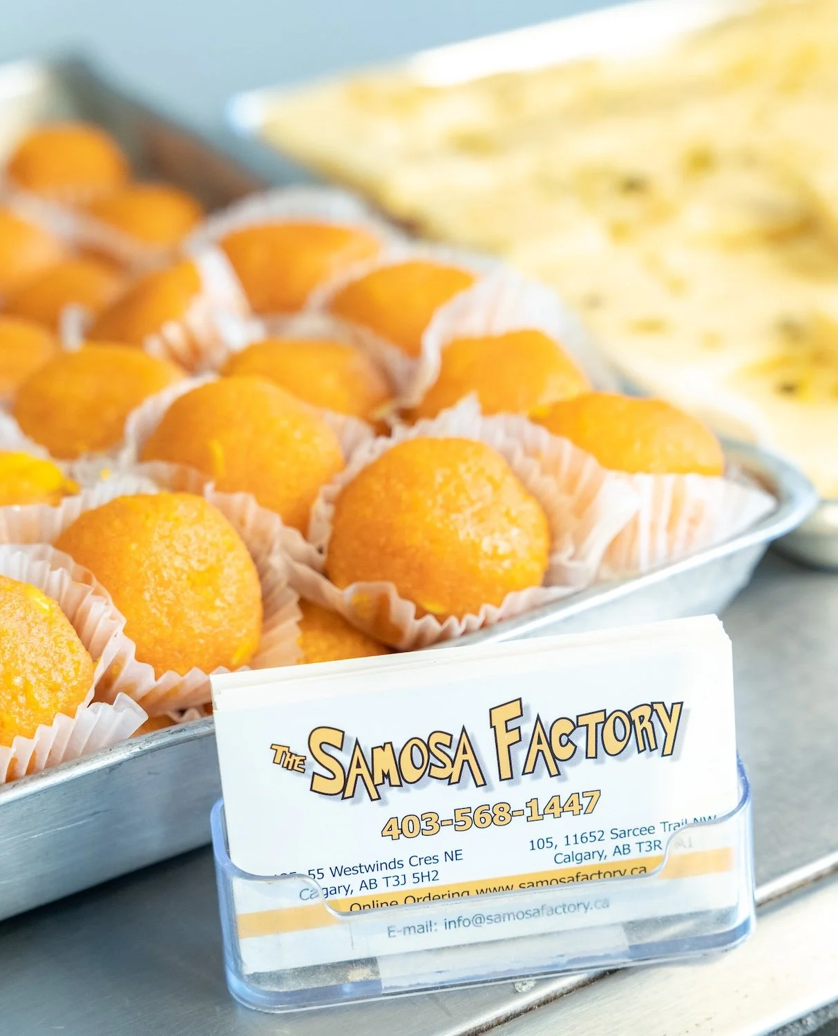 Tray of orange-colored Indian desserts in white paper cups at Samosa Factory with a printed business card in front.