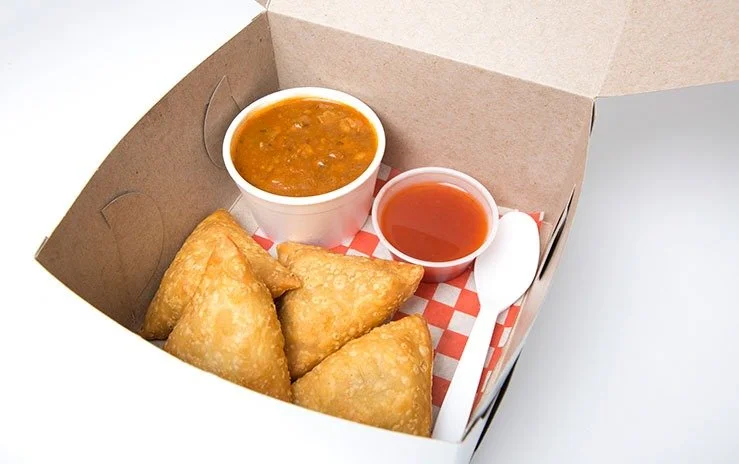 Four fried samosas in a takeout lunch box, with cups of dipping sauce and chickpea curry, and a white plastic spoon.