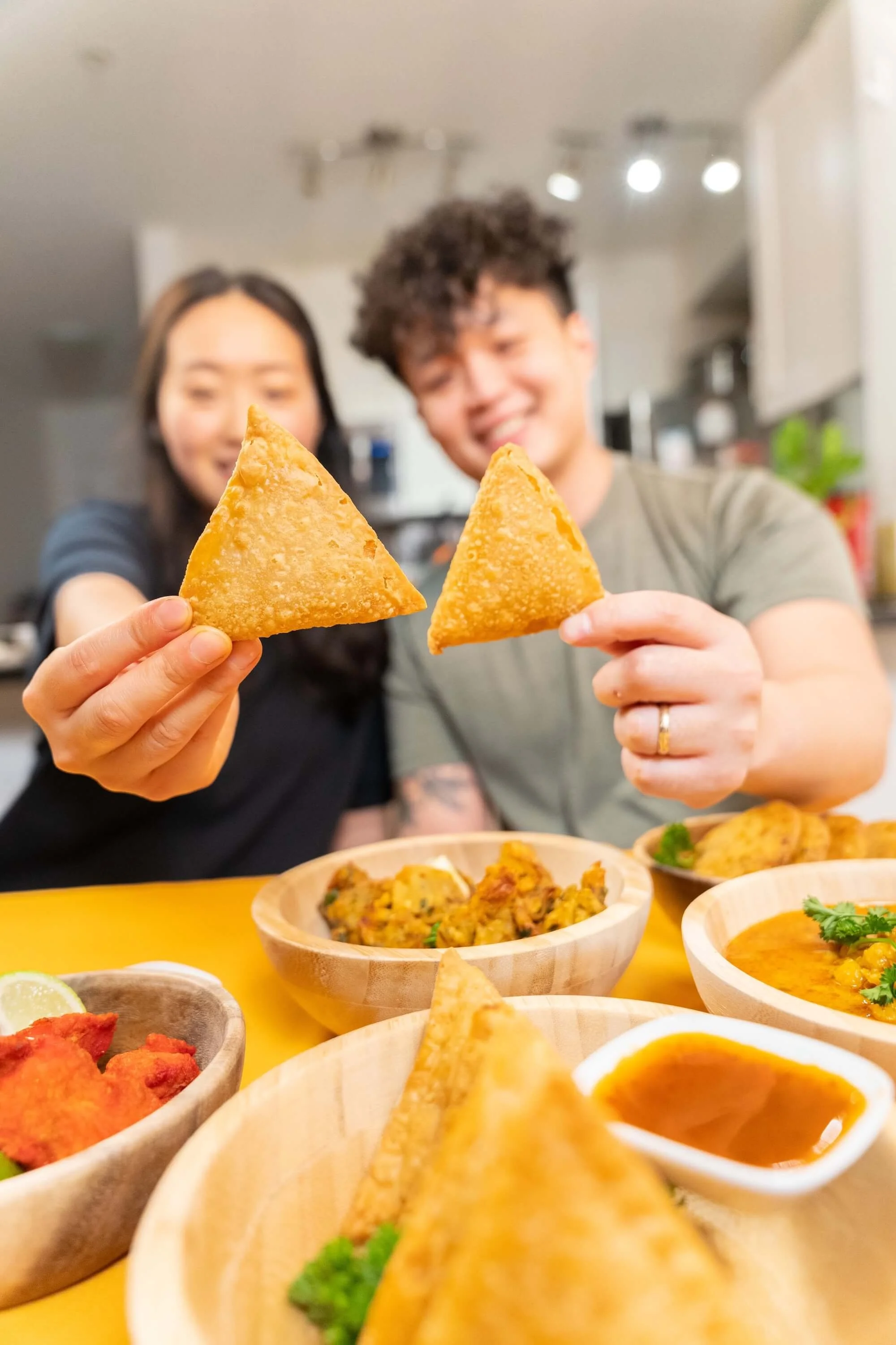 A man and woman holding up two samosas with a variety of Indian dishes and dip on a yellow table.