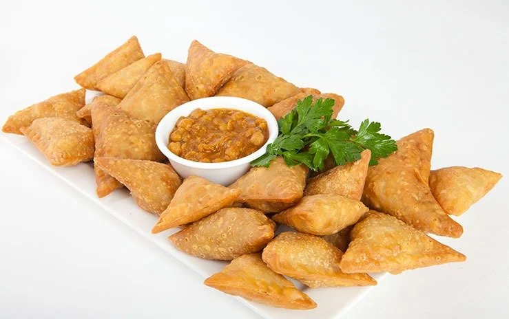 A white rectangular plate with fried samosas, a bowl of chickpea curry, and a sprig of cilantro on top.