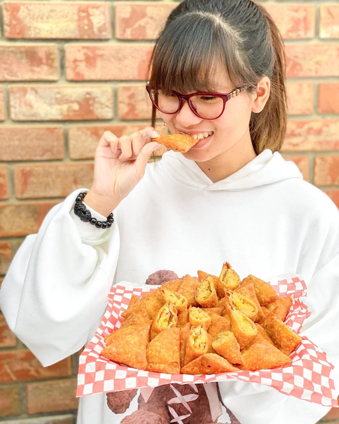 Young woman with glasses holding a basket of fried samosas, one pastry near her mouth.