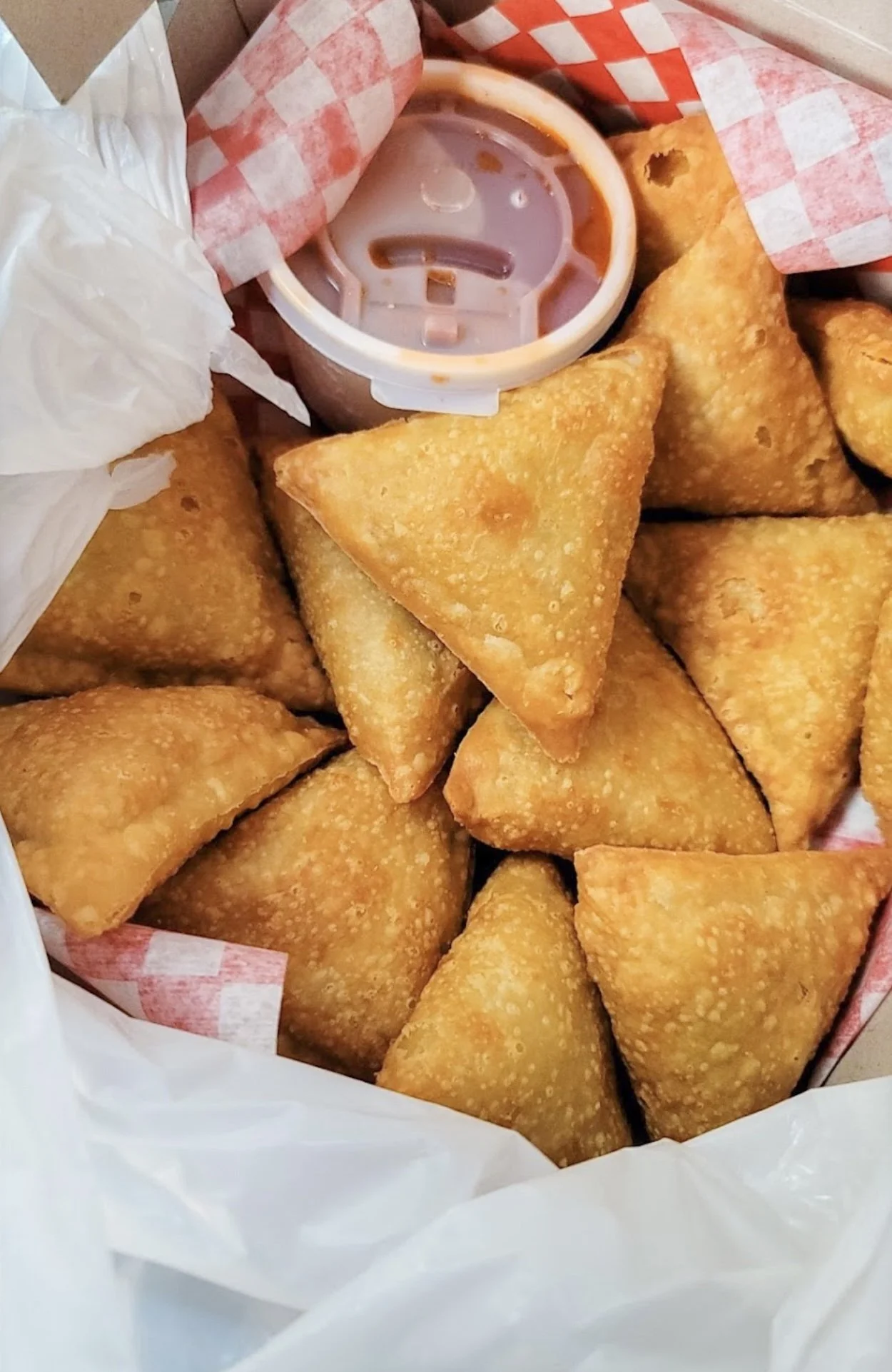 A basket of golden-brown fried samosas with a small container of dipping sauce on top, lined with red and white checkered paper.