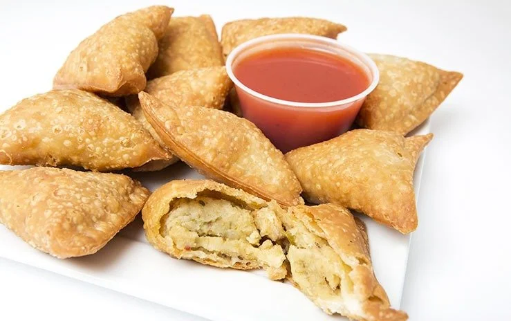 Plate of nine triangular fried samosas with a cup of red tamarind chutney in the center, one dumpling cut open showing the filling inside.