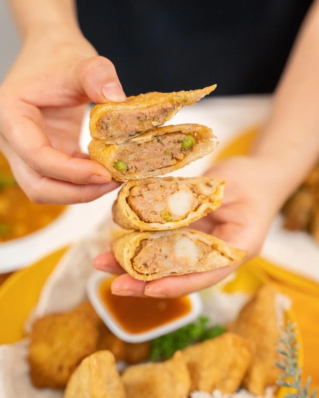 Close-up of a hand holding a samosa with a meat filling and green peas, with additional egg rolls and dipping sauces in the background.