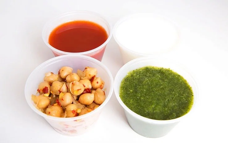 Four small white bowls filled with different East Indian sauces and chickpeas. The sauces include red, white, and green.