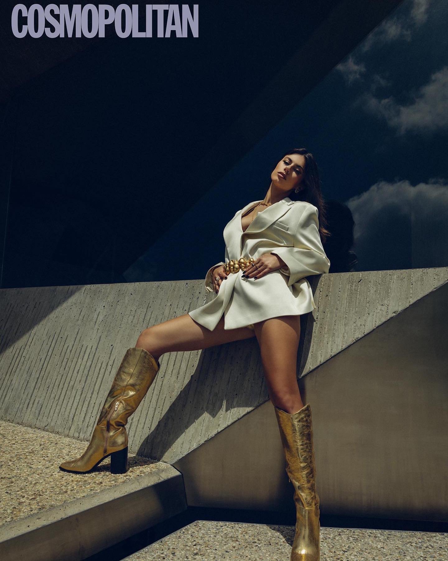 A woman wearing a white blazer, gold knee-high boots, and jewelry, posing against a modern concrete and glass building with a dark sky in the background.