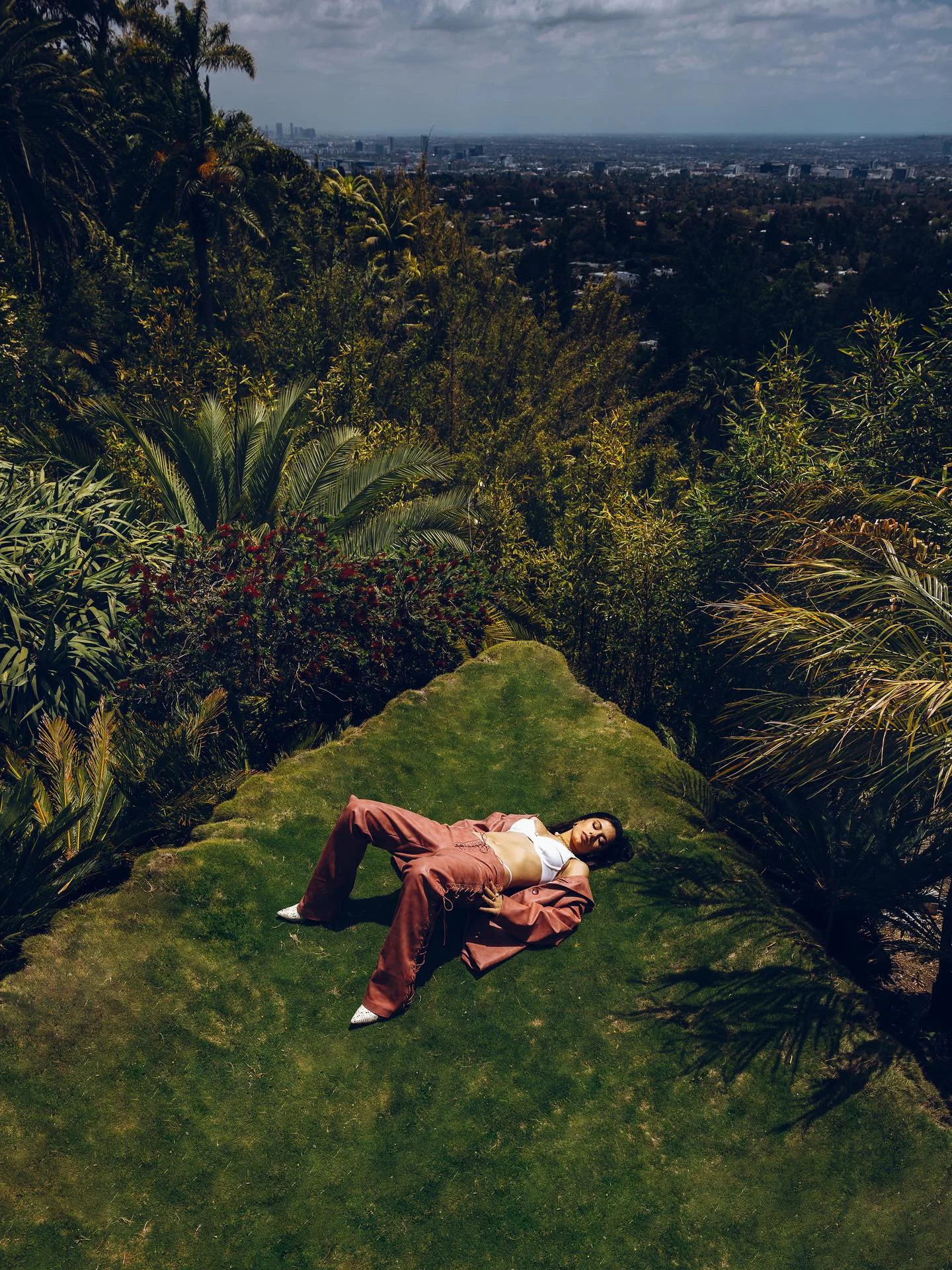 Woman in a white top and brown pants lying on a grassy hill, surrounded by greenery, with a city skyline in the distance under a cloudy sky.