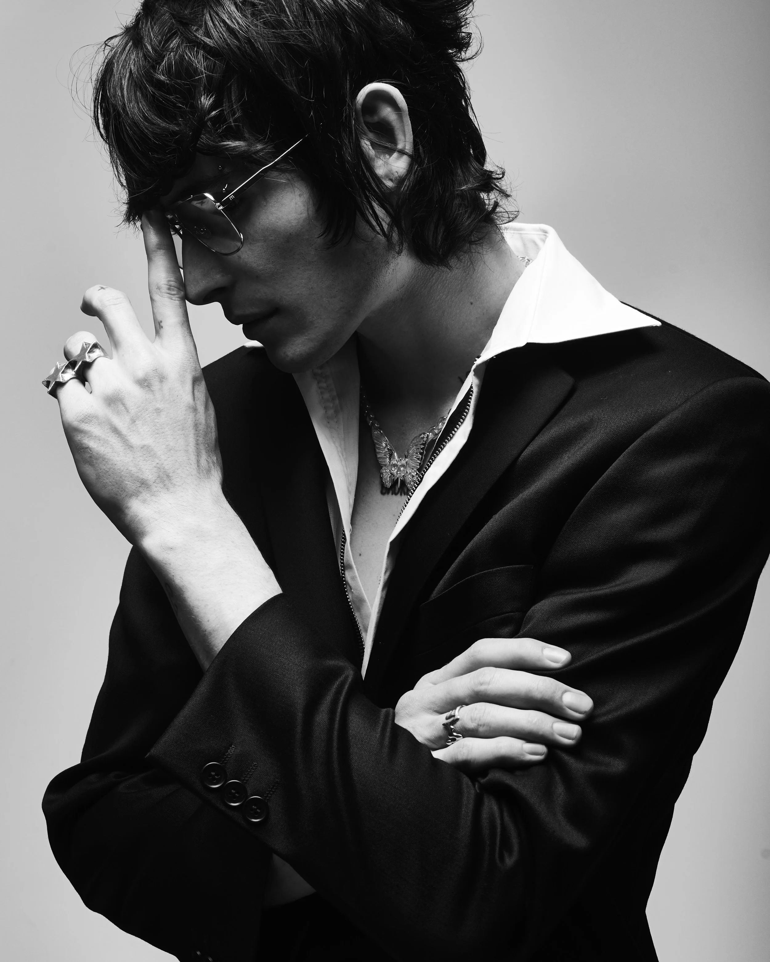 A black-and-white portrait of a young man with wavy hair, wearing sunglasses, a suit jacket, and a white shirt. He is touching his forehead with one hand and has rings on his fingers, along with necklaces around his neck.