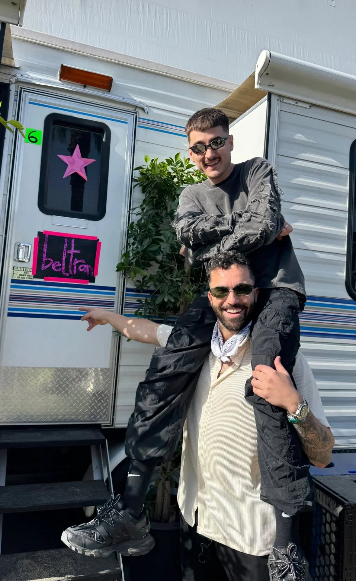 Two men, one giving the other a piggyback ride in front of a trailer door. Both men are wearing sunglasses and smiling. The trailer has a pink star decoration and a sign that says 'beltra' with pink and black colors.