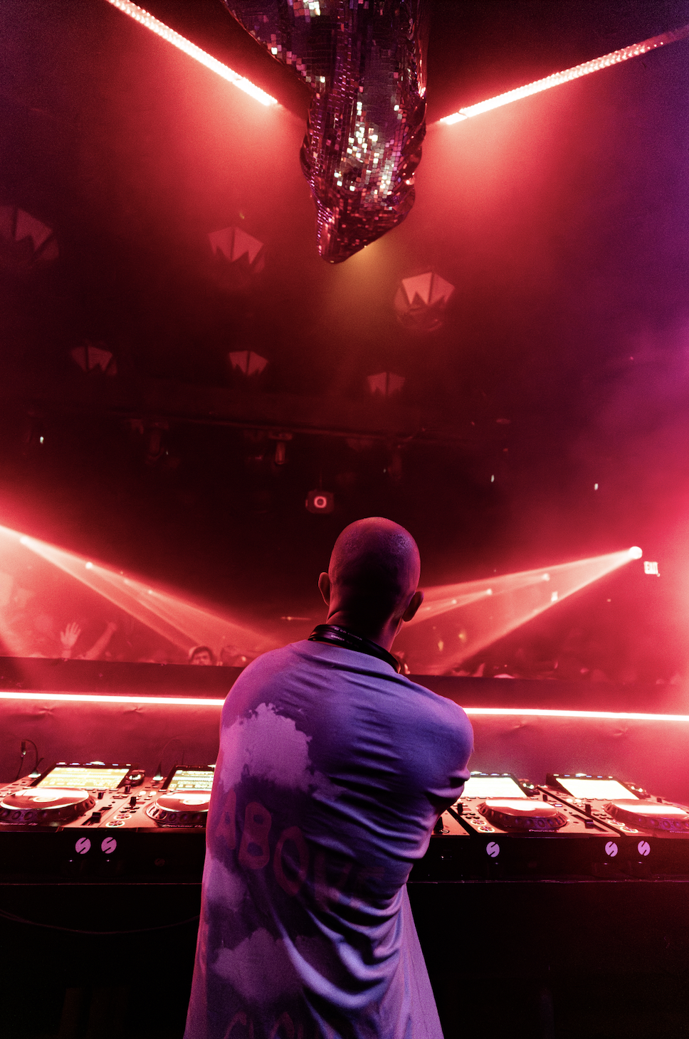 A DJ stands in front of a DJ booth in a nightclub with red and pink lighting, a mirrored disco ball on the ceiling, and an audience with raised hands visible in the background.
