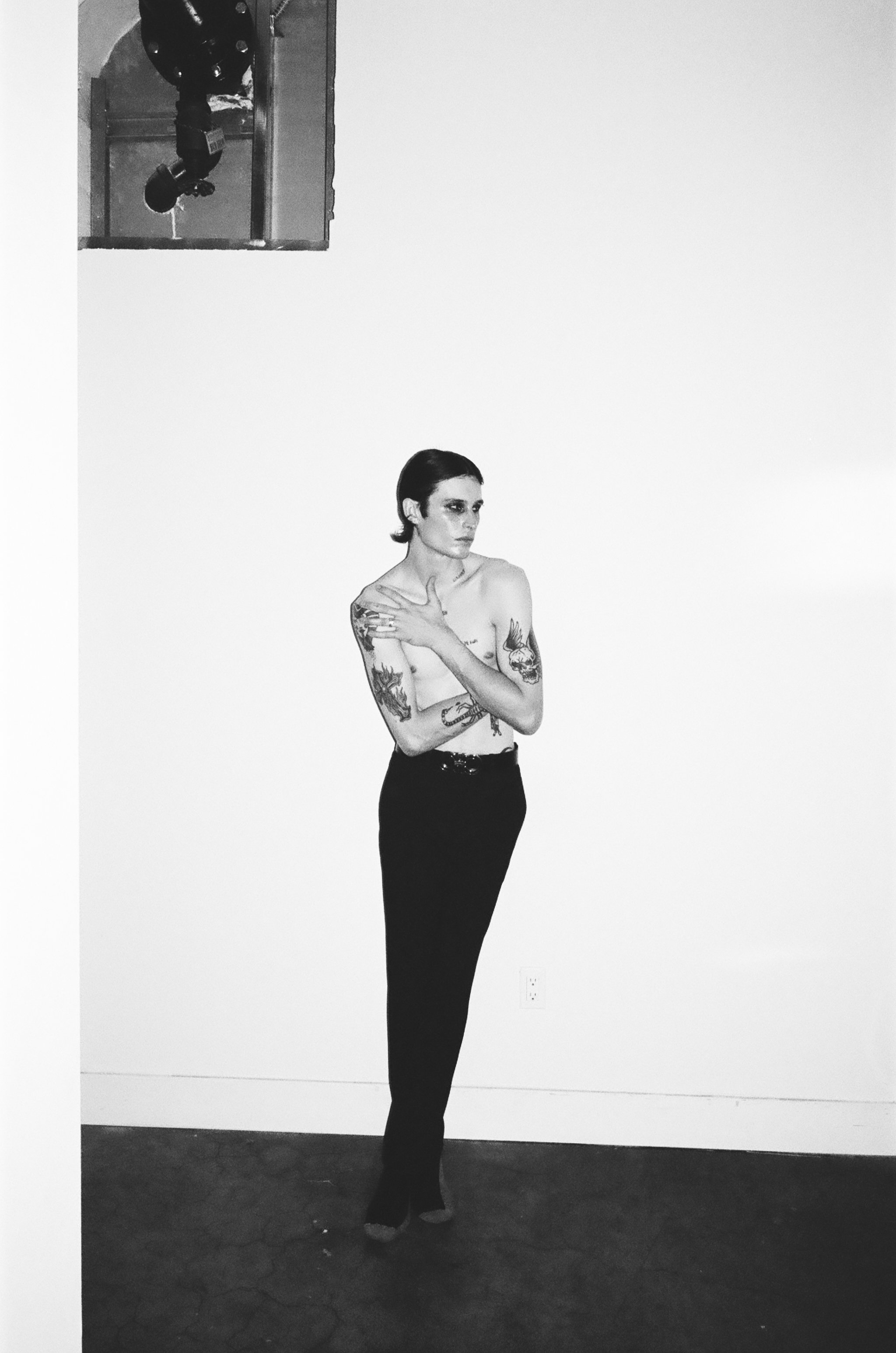 A black-and-white photograph of a tattooed woman with short dark hair, standing against a plain white wall, wearing high-waisted black pants, with an inset photo showing a person in a different setting at the top left corner.