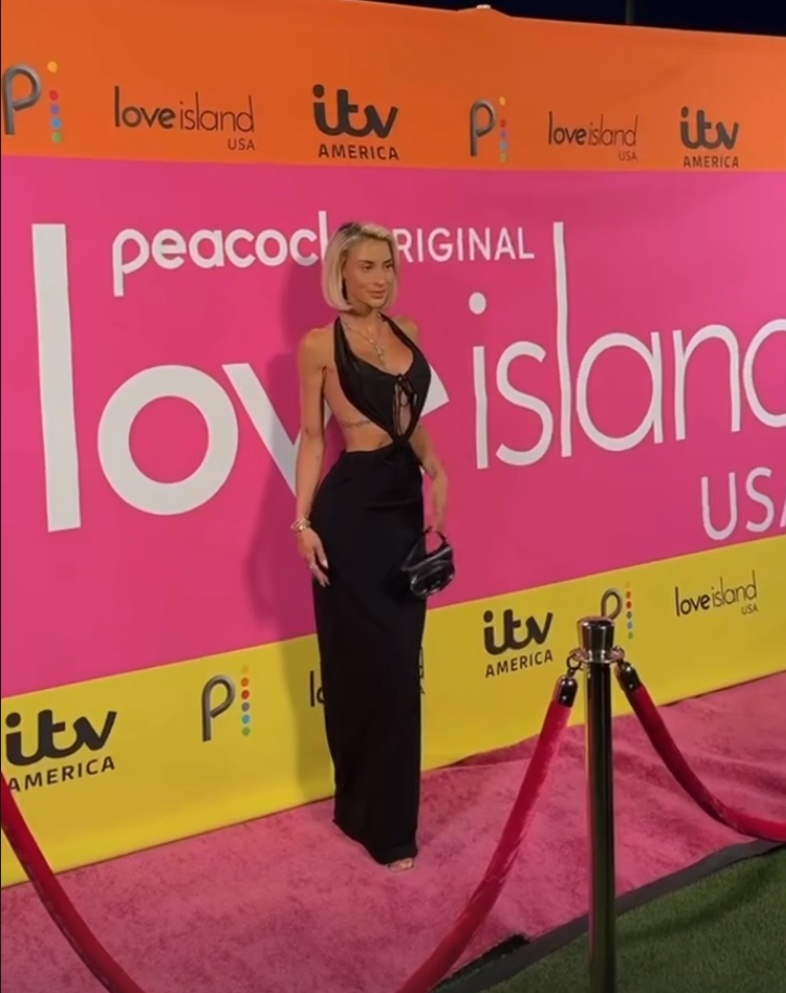 A woman in a black outfit standing on a pink carpet with a pink and yellow backdrop behind her that reads 'Love Island USA' and 'ITV America.'
