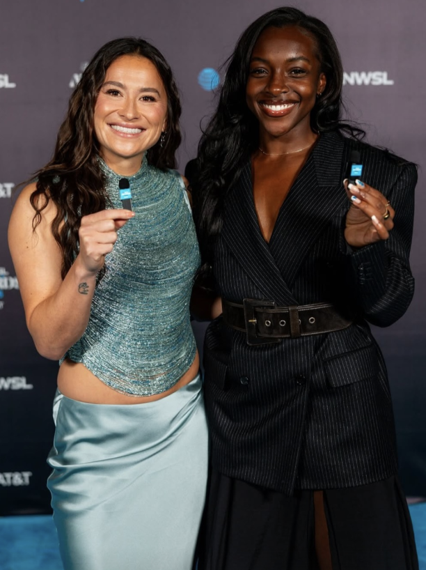 Two women smiling and holding small award statues at an event, standing in front of a backdrop with logos.