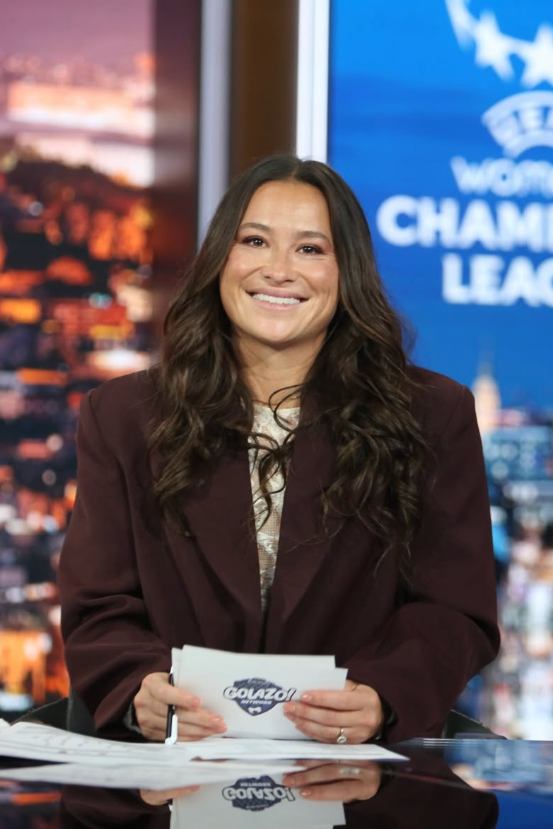 A woman with long brown wavy hair smiling and holding notes at a news desk, with a sports league logo on the background screen.