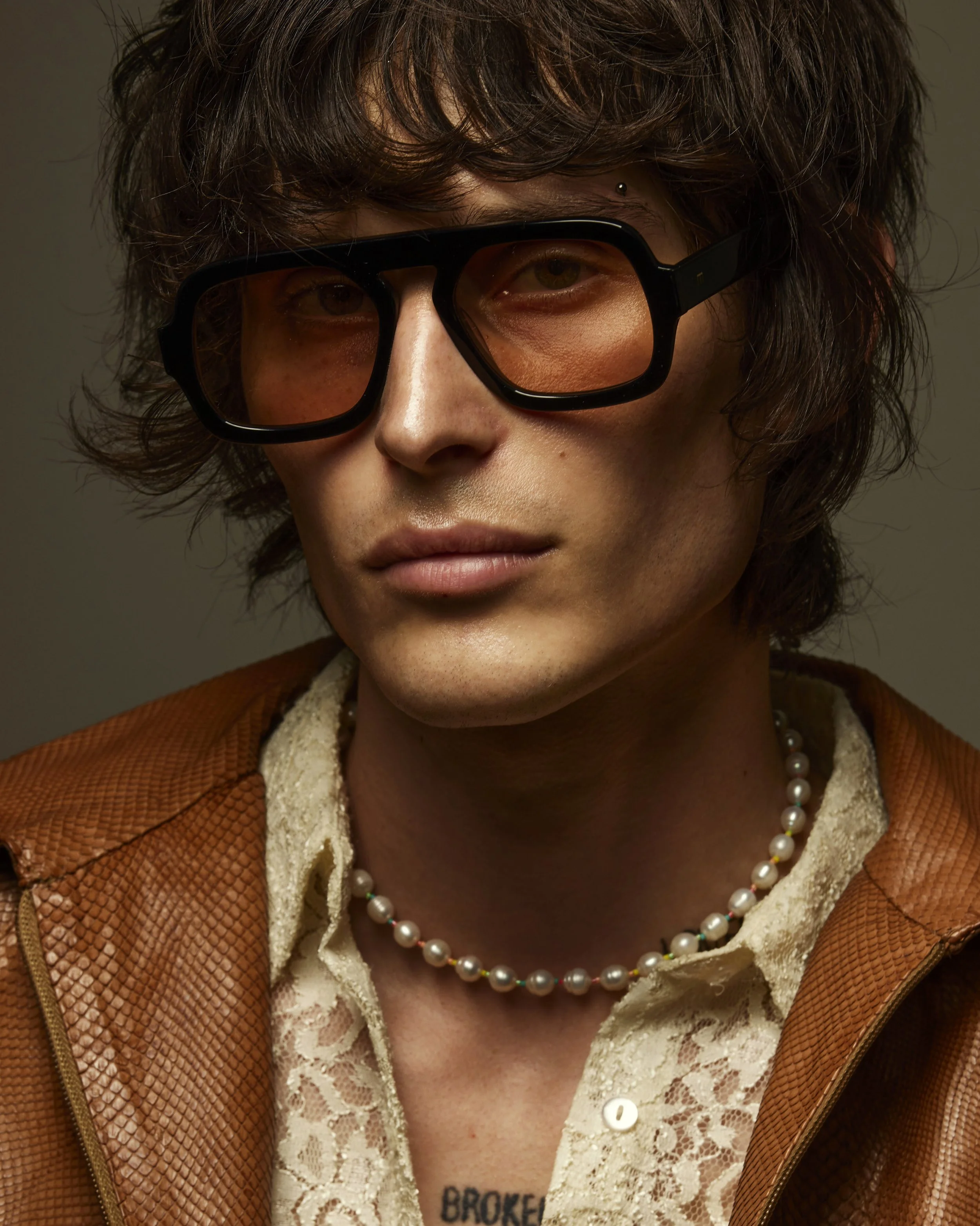 Close-up of a young person with wavy brown hair wearing large black glasses with orange-tinted lenses, a pearl necklace, a lace shirt, and a brown leather jacket.