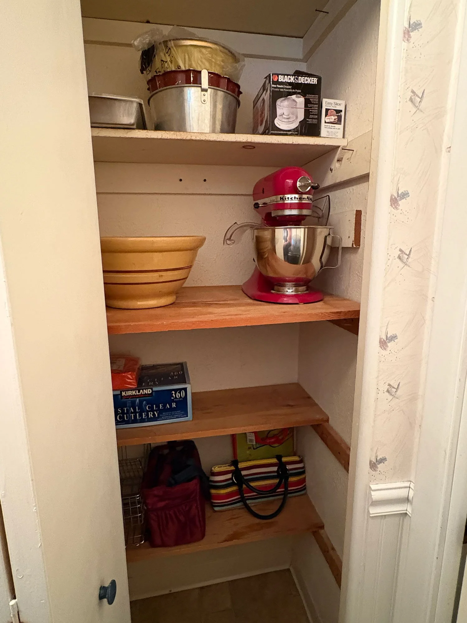 Empty kitchen cabinet with three shelves holding various kitchen items including a bowl, boxes, a small candy jar, a stand mixer, a bag, and other miscellaneous items.