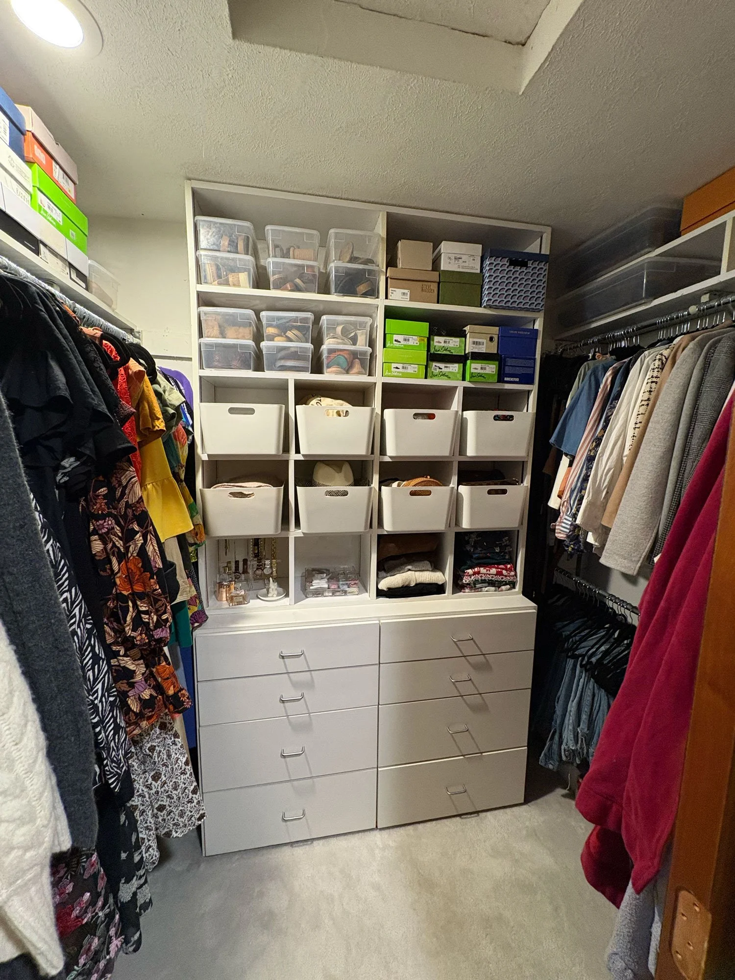 A walk-in closet with clothes hanging on both sides, white storage shelves with shoes and boxes in the center, and drawers at the bottom.