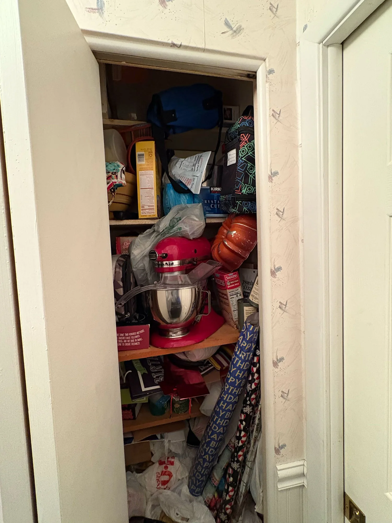 A small, cluttered closet filled with various items including a gift bag, wrapping paper rolls, a plastic bag, a red stand mixer, and food packaging on shelves.