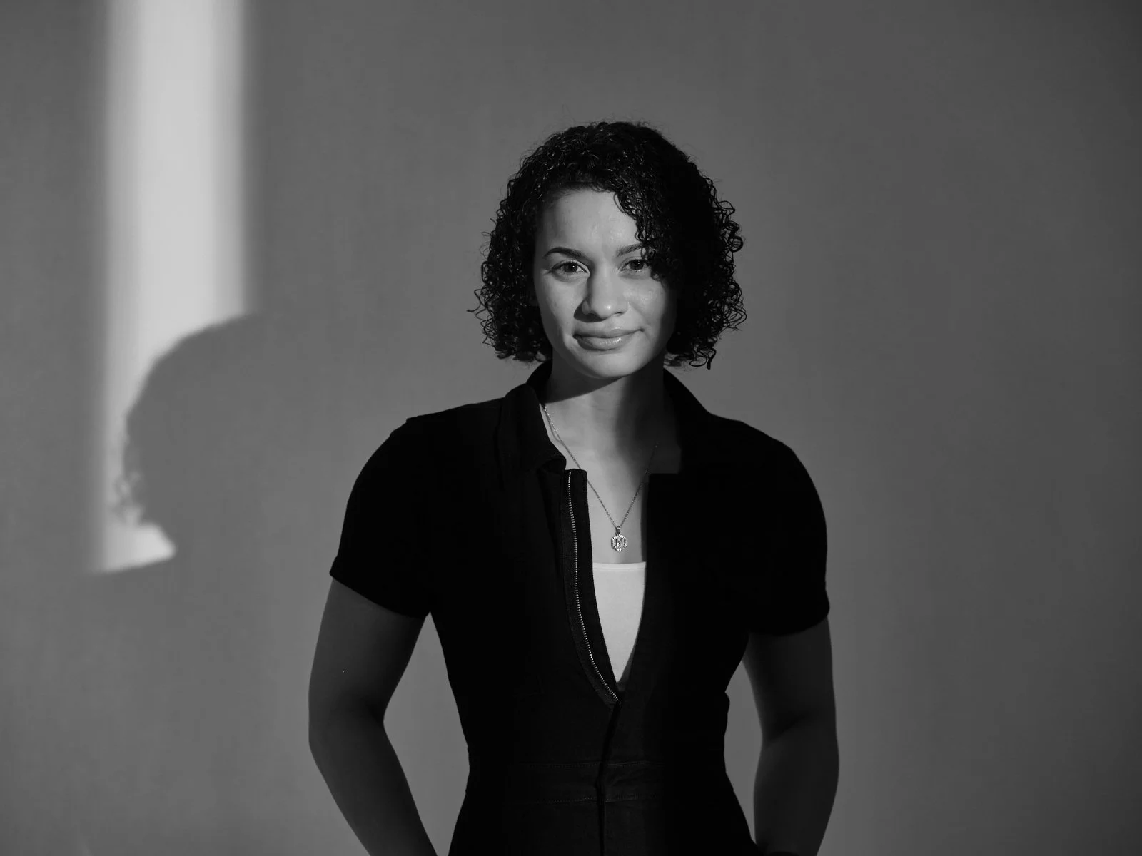 Black-and-white portrait of a woman with short curly hair, wearing a dark top and necklace, standing against a plain background with soft, directional lighting and a calm, confident expression.