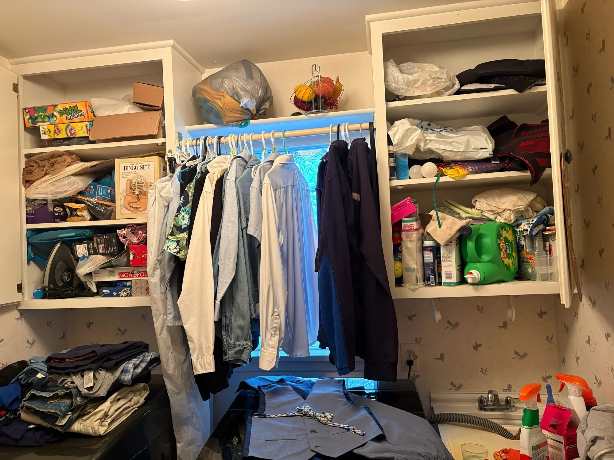 Cluttered laundry room with open cabinets, clothes hanging on a rack, and various household items on shelves.