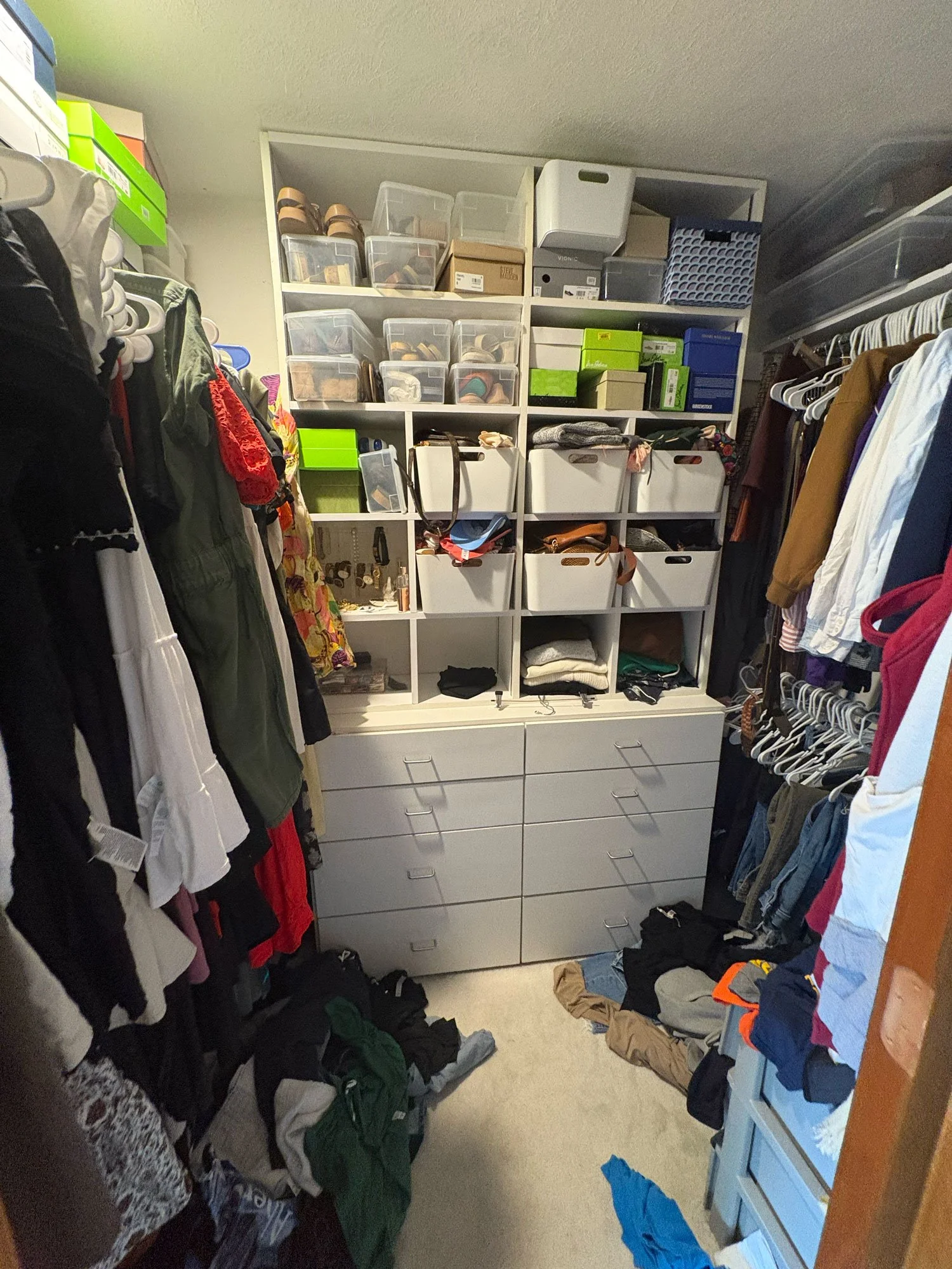 A walk-in closet with hanging clothes on both sides, shoes stored on shelves above, and some clothes and items on the floor.