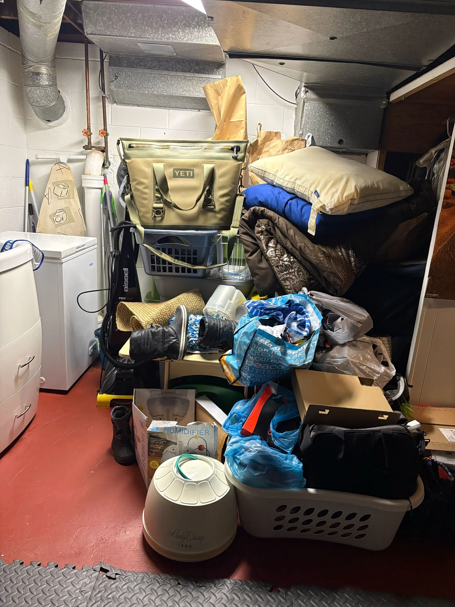 A cluttered storage room with various household items, including laundry baskets, a humidifier, bedding, and cleaning supplies, all stacked and scattered.