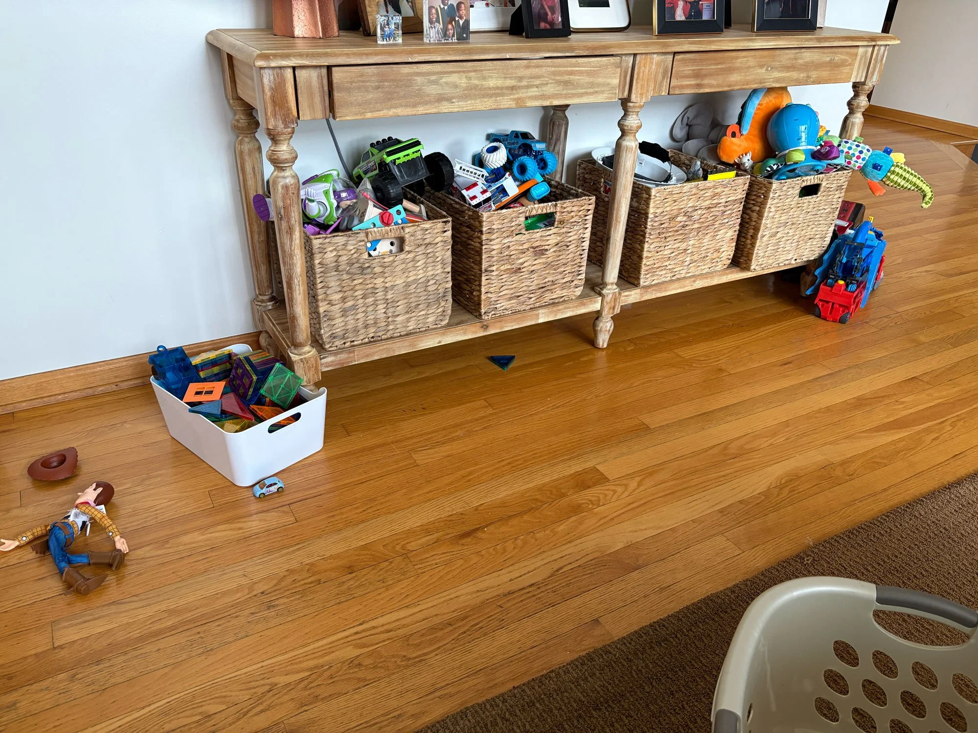 A wooden console table with four wicker bins filled with children's toys like cars, figures, and building blocks. Toys are scattered on the wooden floor in front.
