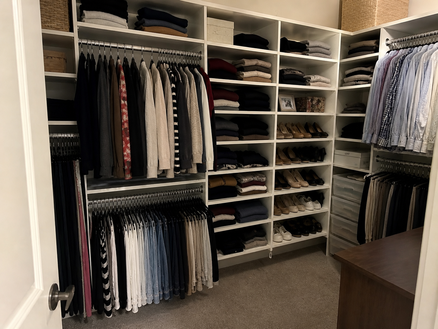 Walk-in closet with white built-in shelving, neatly hung clothing, folded sweaters, and organized shoes. Soft, natural lighting and a slightly lived-in feel create a realistic, tidy but not overly staged space.