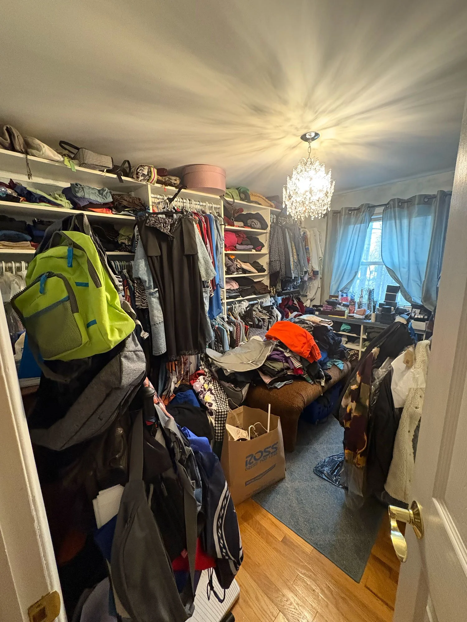 A cluttered bedroom with shelves filled with clothes, bags, and boxes. There are hanging clothes, a chair with clothes draped over it, a bag on the floor, and a window with curtains. A chandelier hangs from the ceiling.