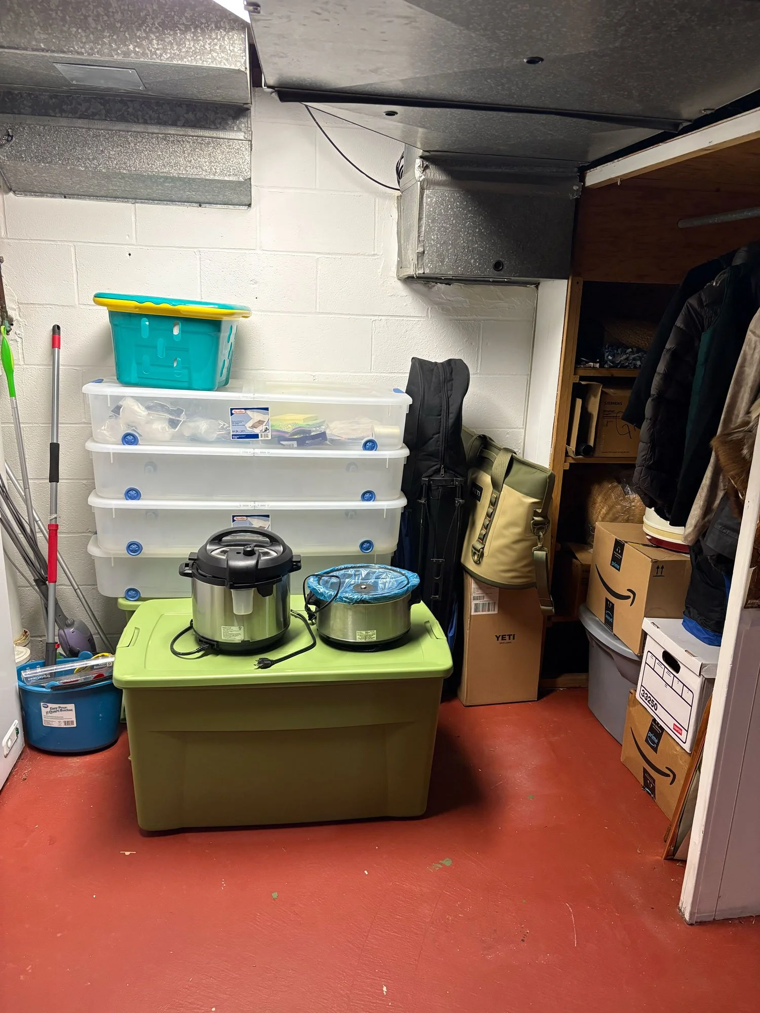 A small indoor storage room with red flooring, white painted cinder block wall, and a green storage bin on the floor. On top of a white plastic drawer storage unit, there is a teal plastic basket. Next to the storage unit, there are various bags and 