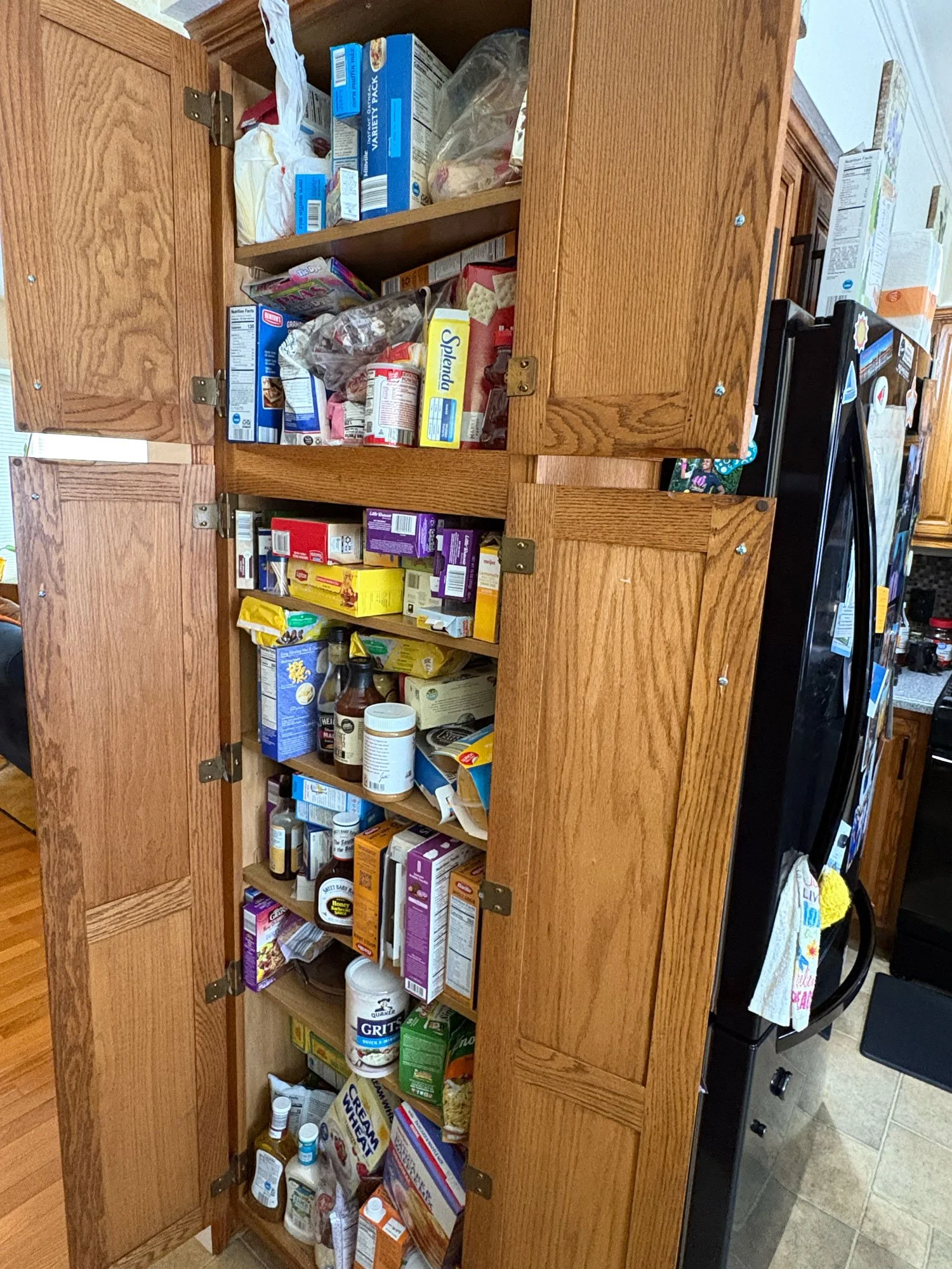 Open kitchen cabinet filled with various groceries, including cereal, canned goods, snacks, and bottles of condiments. Part of a black shelf with a door towel hanging on it is visible to the right.
