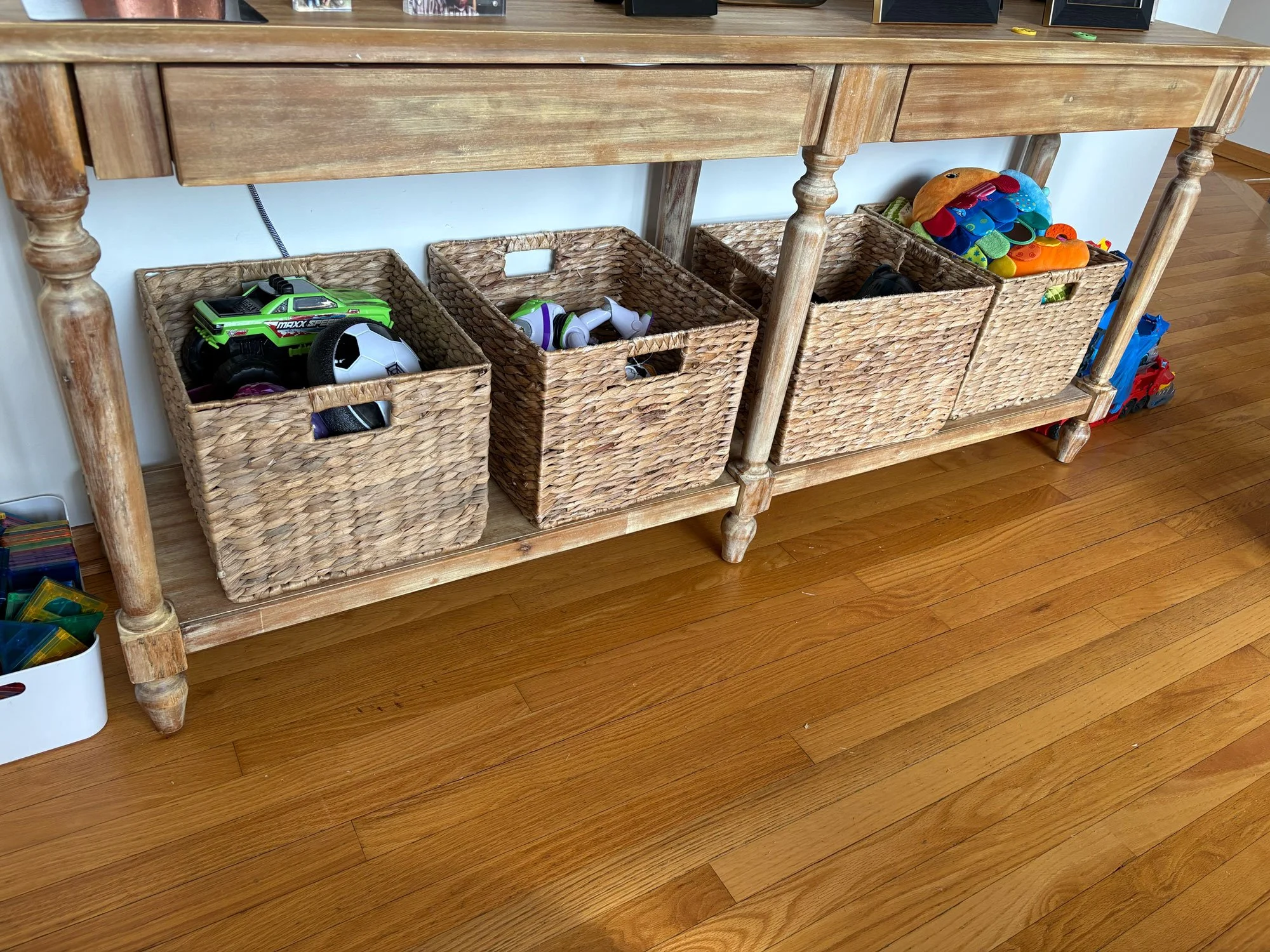 A wooden console table with four woven baskets underneath, filled with children's toys such as cars, a soccer ball, plush toys, and various small play items, located on a hardwood floor.