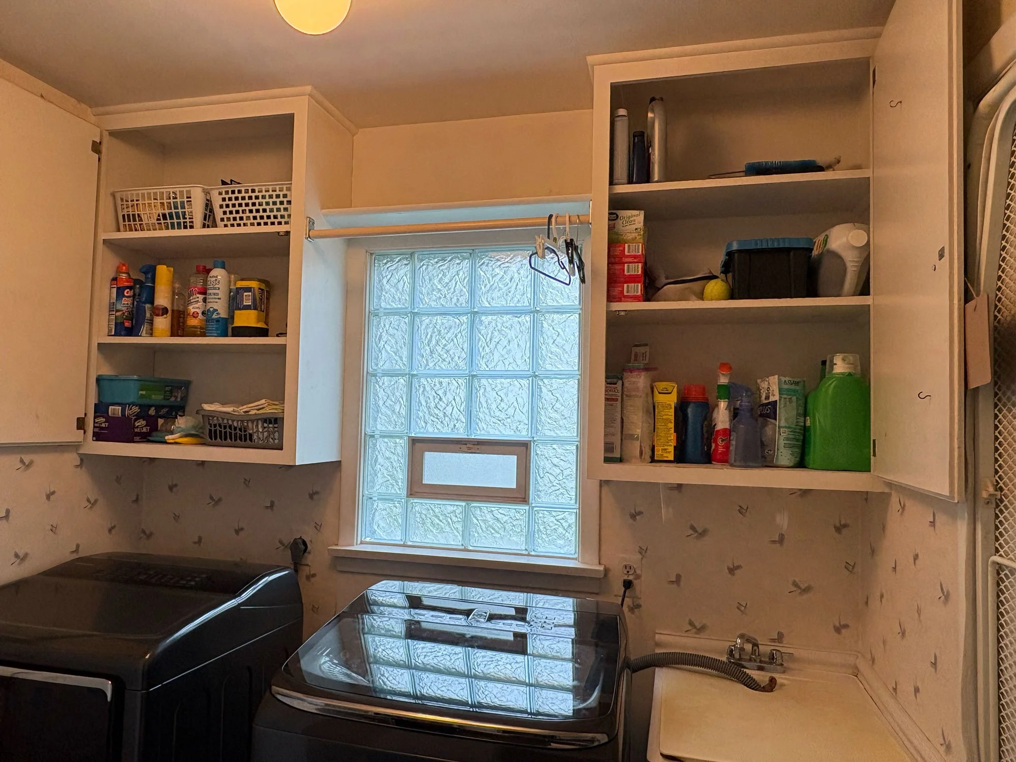 Kitchen with white cabinets, a window with glass blocks, and appliances including a microwave and a washing machine. Items such as boxes, bottles, and laundry supplies are on the shelves.