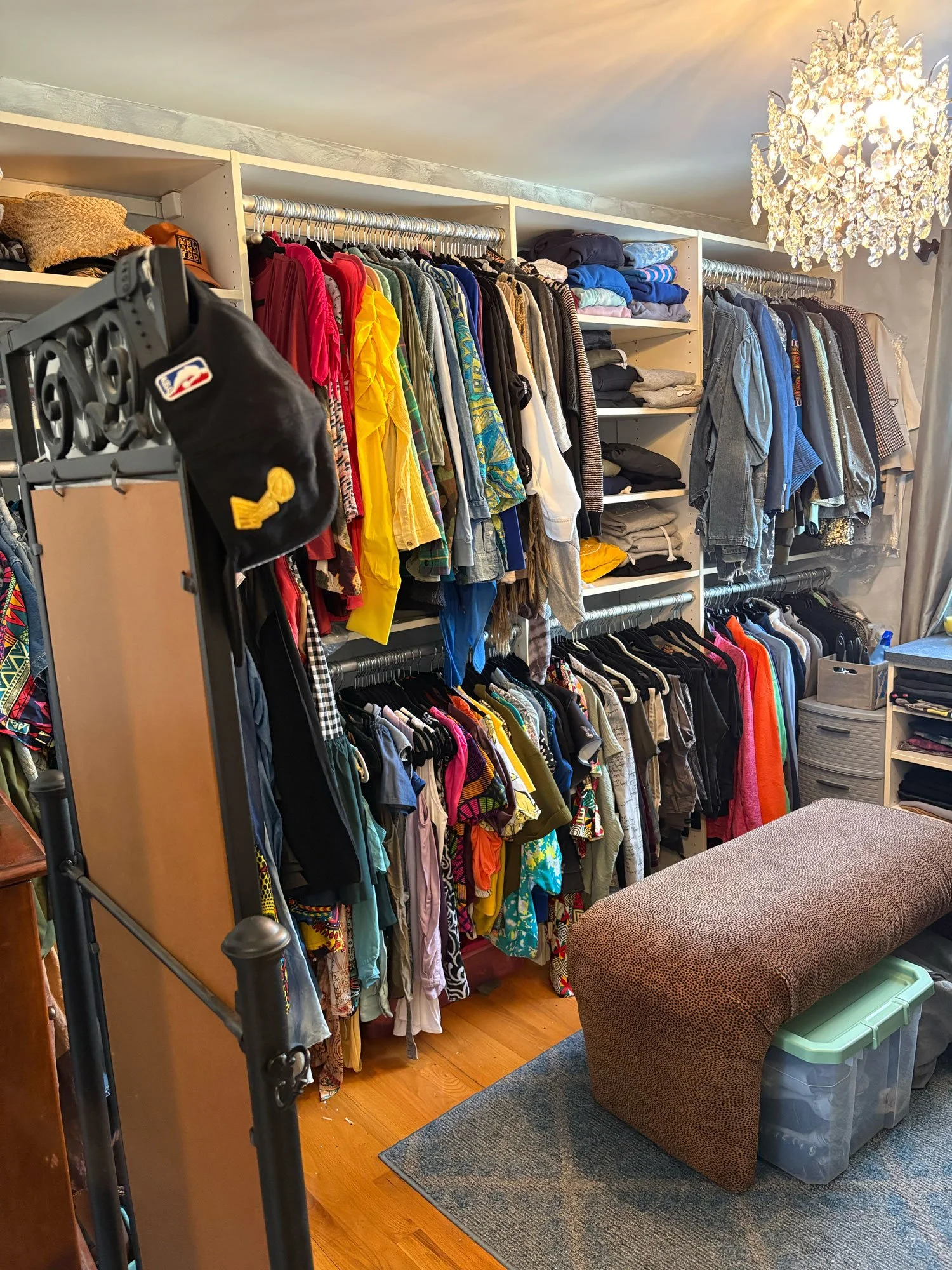 A walk-in closet with multiple shelves and hanging rods filled with clothes, including shirts, jackets, and pants. There is a chandler chandelier hanging from the ceiling, a brown upholstered ottoman, and a grey storage bin on a wooden floor with a s
