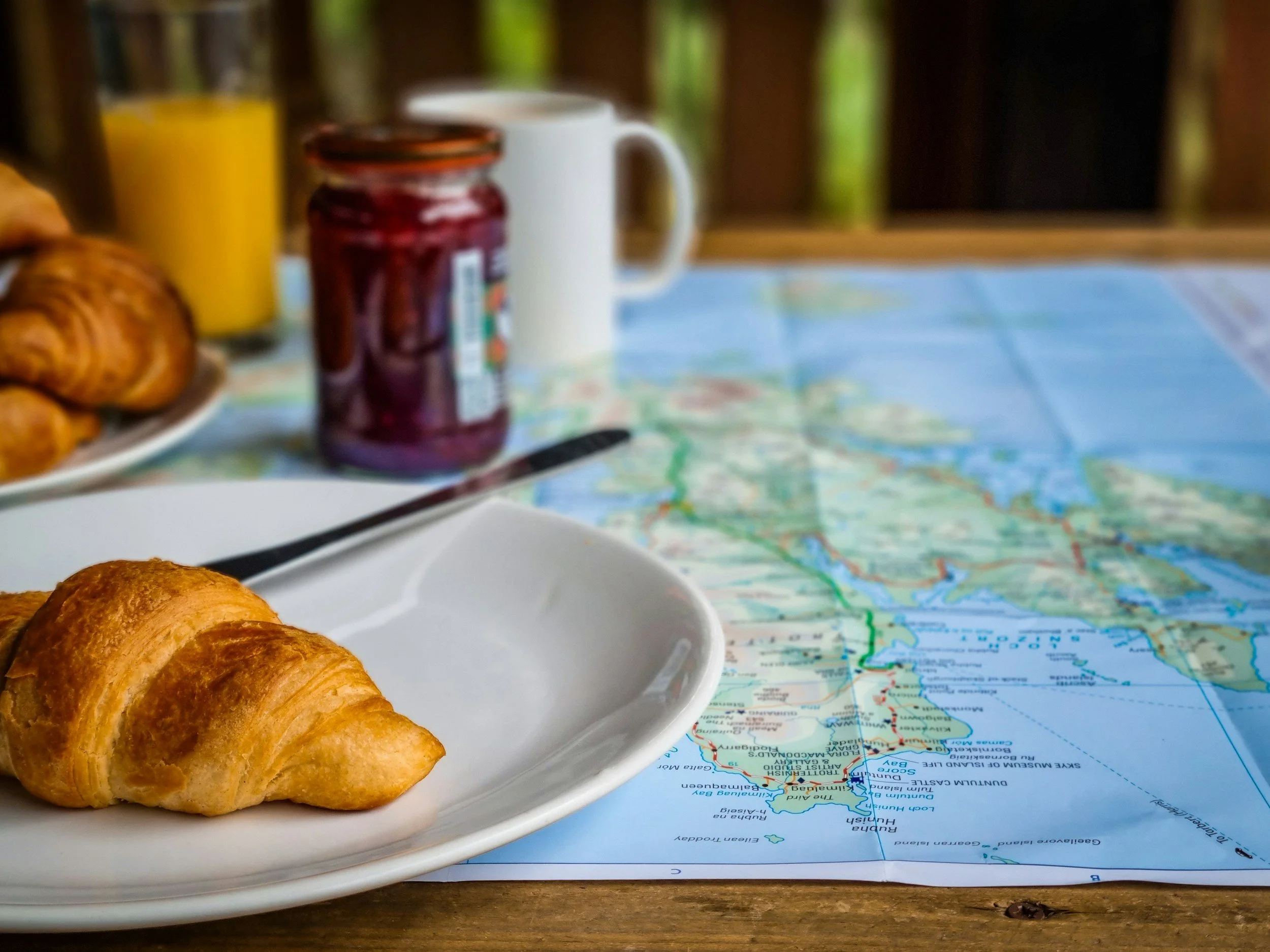 A breakfast scene with a croissant on a white plate, a jar of jam, a cup of coffee, and a map on a table. The background is a wooden fence and trees.