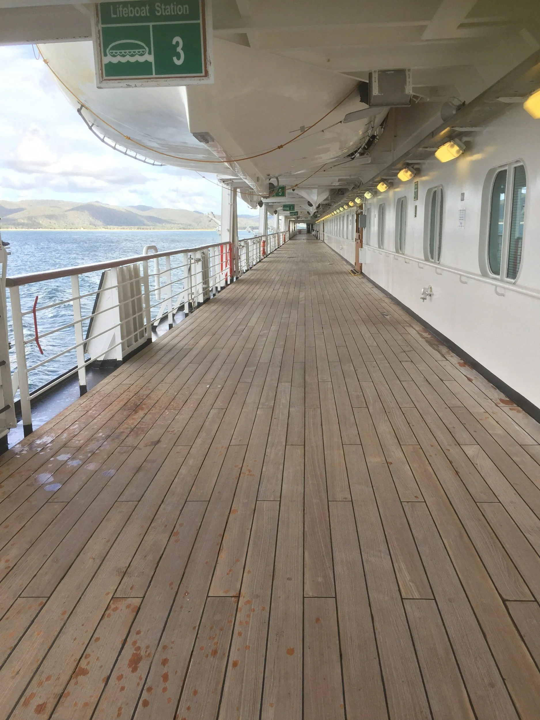 View of a ship's outdoor deck with wooden planks, yellow lights along the side, and a green sign indicating a lifeboat station number 3. The ocean and a mountain range are visible in the background.