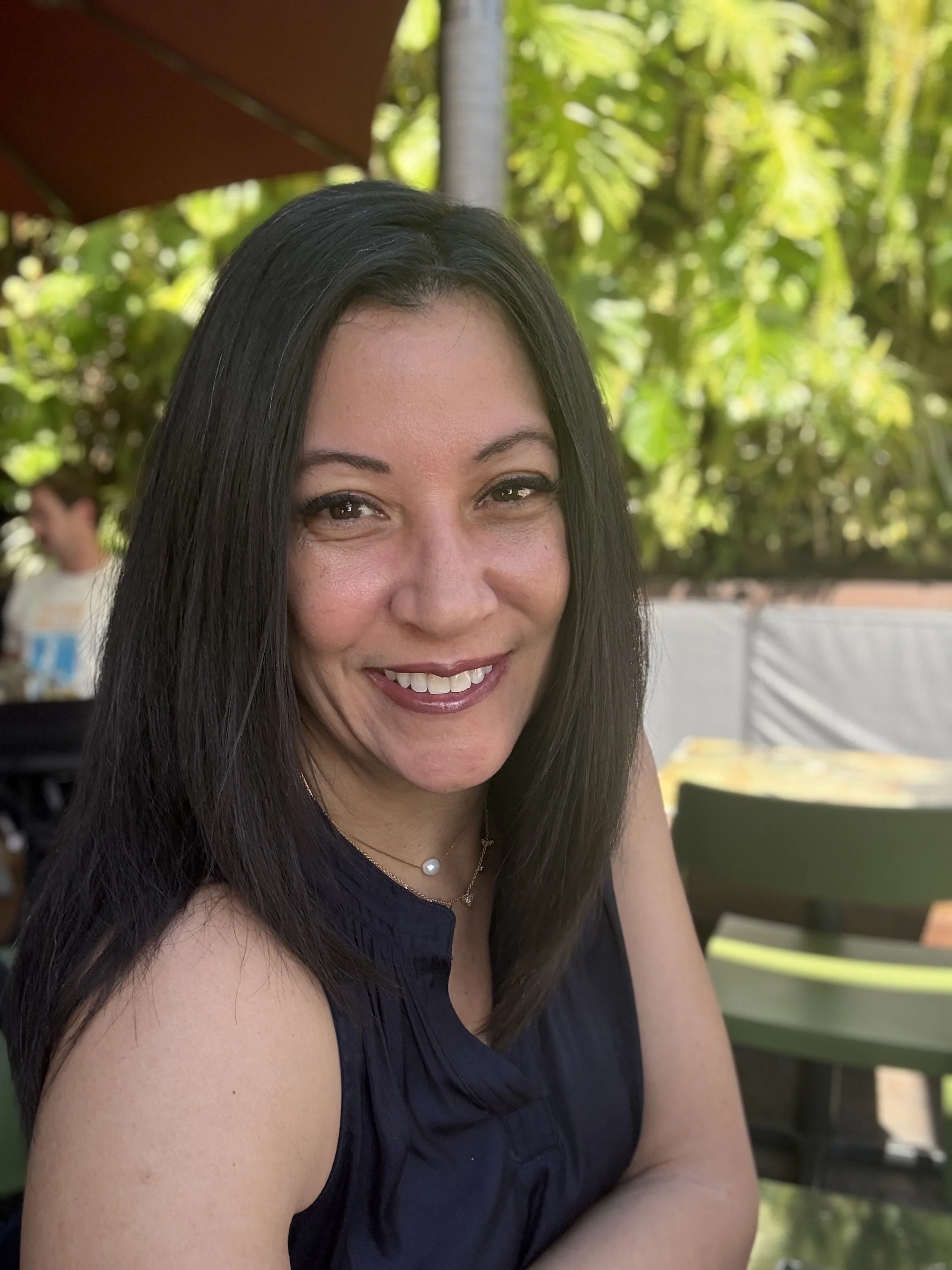 A woman with long dark hair smiling, sitting outdoors at a table with green chairs and surrounded by lush greenery.