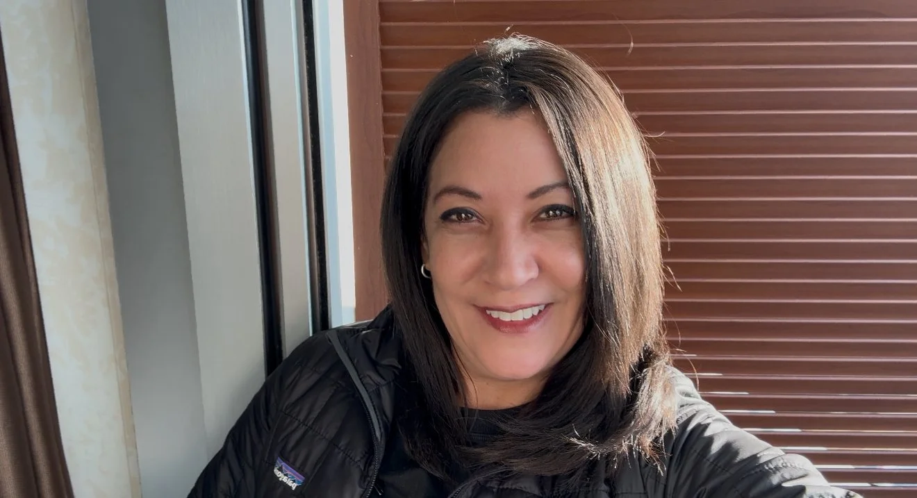 A woman with shoulder-length dark hair smiling at the camera, wearing a black jacket, standing indoors near wooden blinds and a window.