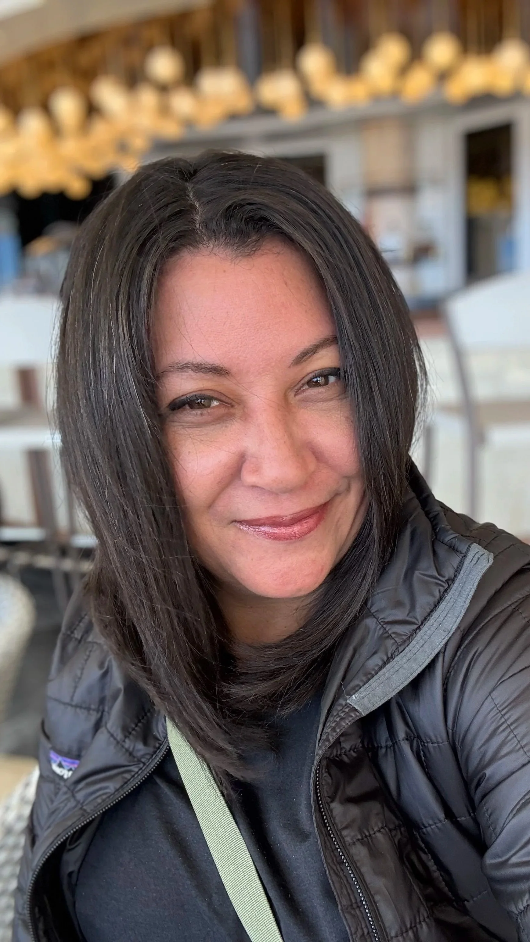 A woman with shoulder-length black hair taking a selfie, smiling at the camera. She is wearing a black jacket and a black shirt with a lime green strap around her neck. The background shows a blurred modern indoor setting.
