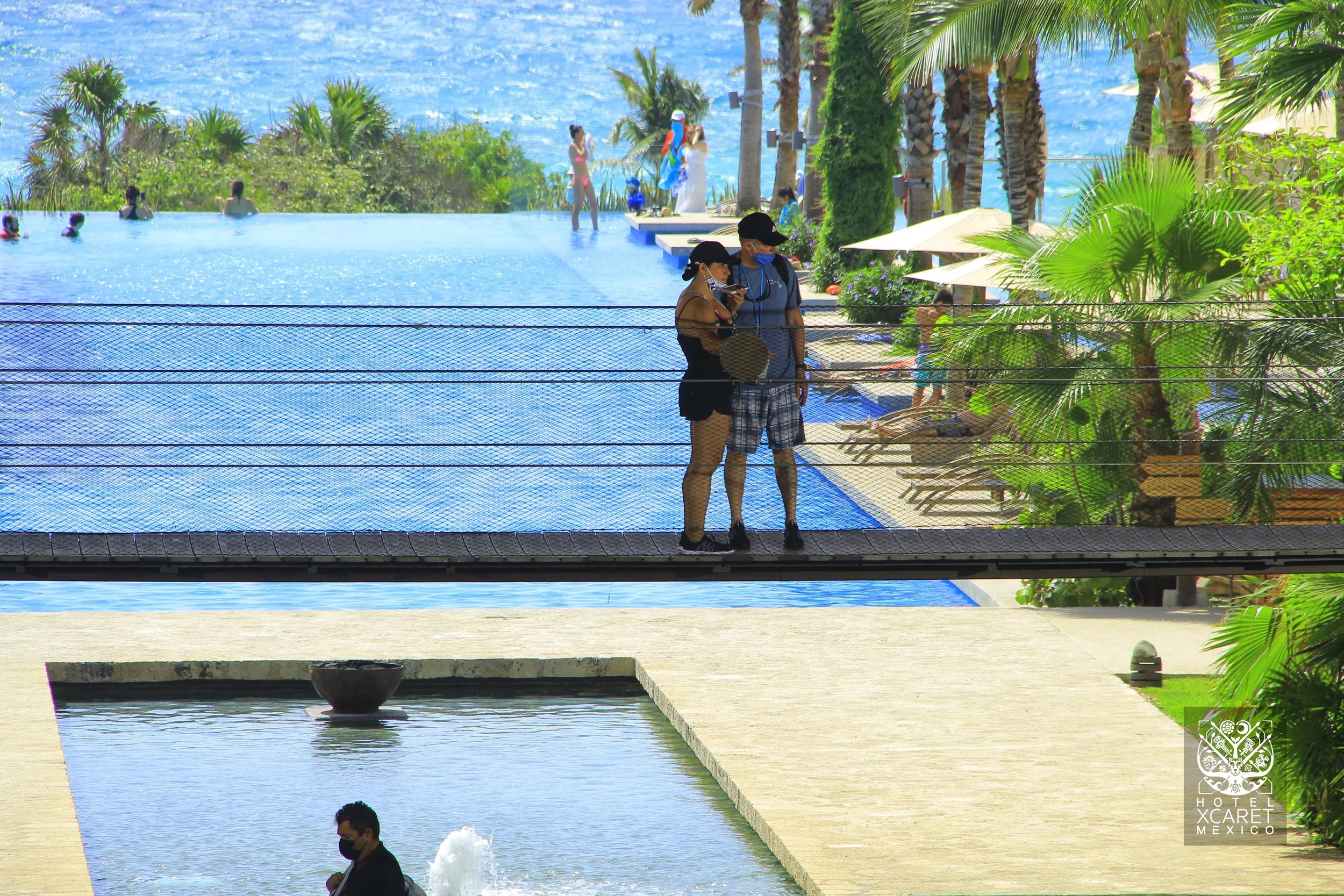 Couple standing on a bridge overlooking an infinity pool at a tropical resort with palm trees, lounge chairs, and people swimming in the pool. Ocean in the background.