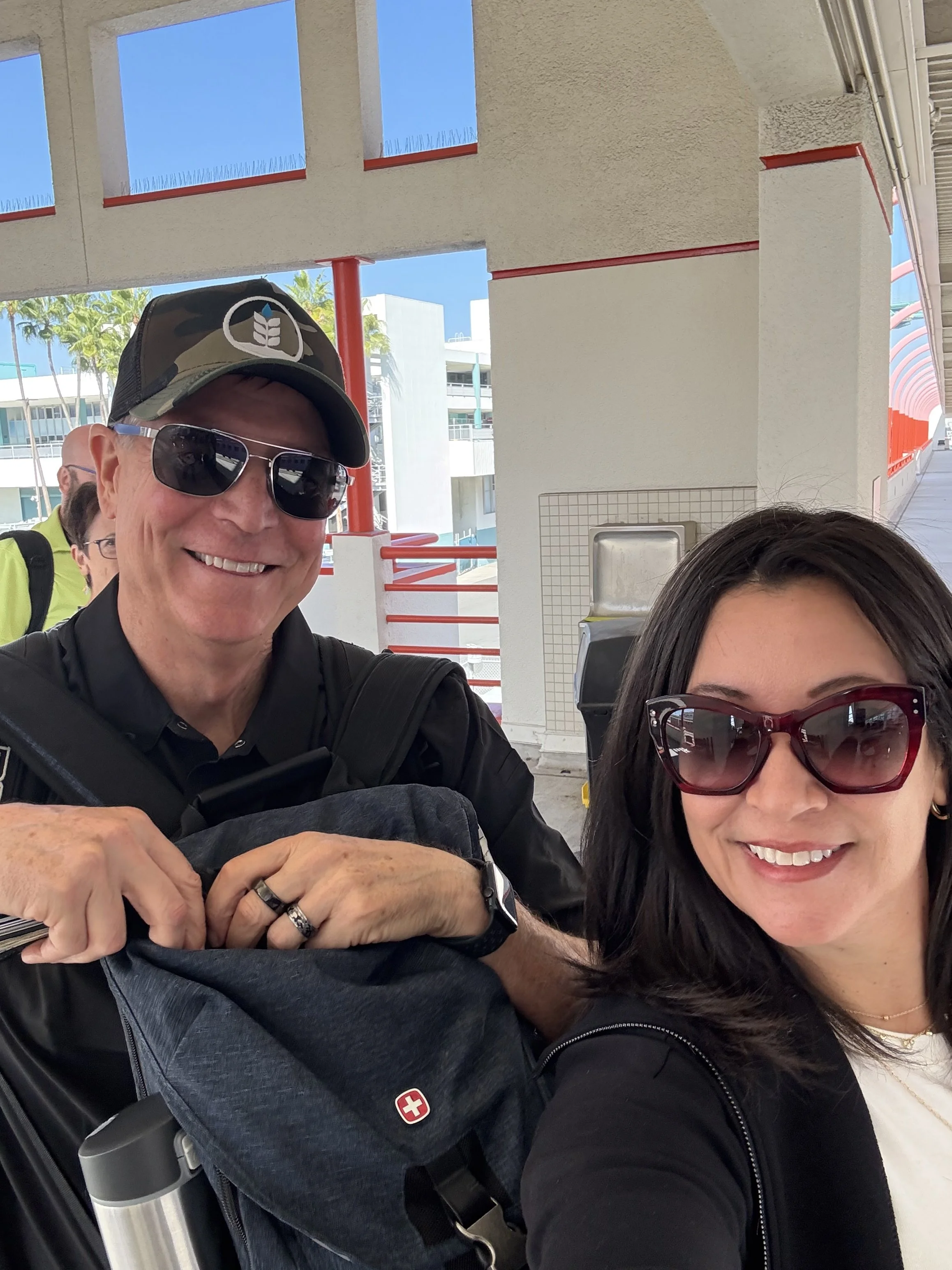 Two smiling people taking a selfie at an outdoor location, with a man wearing sunglasses, a camouflage hat, and a black shirt, and a woman with dark hair, wearing sunglasses, a white top, and a black jacket.